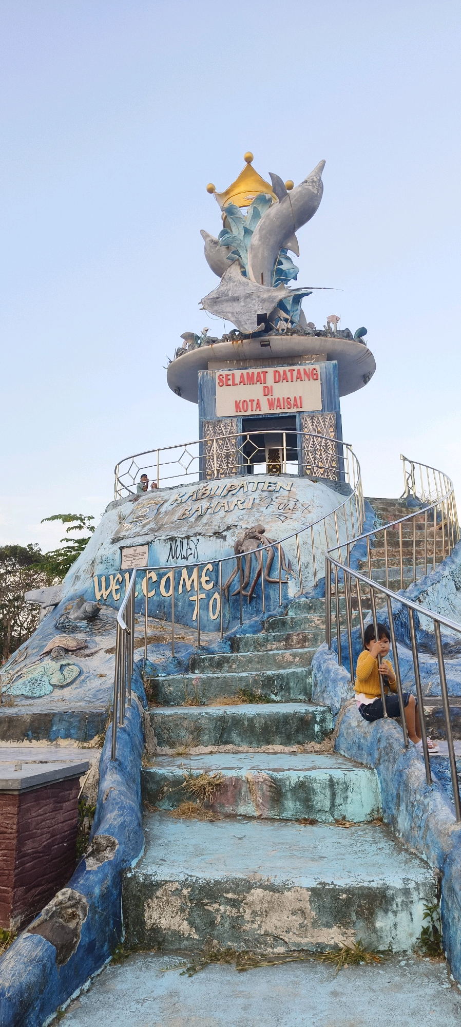 The image depicts a statue located in Waisai, Indonesia. The statue is blue and white, with several dolphins and a crown at the top. There is a sign that says "SELAMAT DATANG DI KOTA WAISAI," which translates to "Welcome to Waisai City." Below the sign, there are the words "KABUPATEN BAHARI PULAU," meaning "Marine Regency Islands." The statue is situated on a hill with a staircase leading up to it. A young child in a yellow shirt and dark shorts is sitting on the stairs. The environment appears to be outdoors with a clear sky. The statue has artistic carvings of sea creatures on its base, including an octopus and a turtle. The overall scene suggests a tourist attraction or landmark in Waisai.