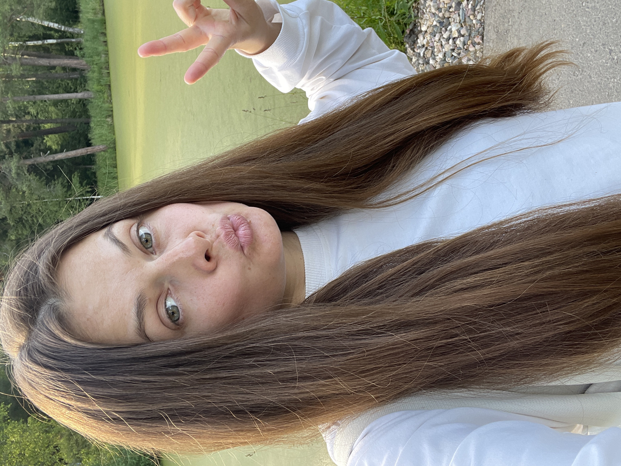 A selfie of a young woman with long, brown hair and green eyes is shown. She is wearing a white long-sleeved shirt. She is puckering her lips and making a peace sign with her right hand. In the background, there is a body of water covered in green algae, surrounded by trees. The location is near the city of Zarechny, Russia.
