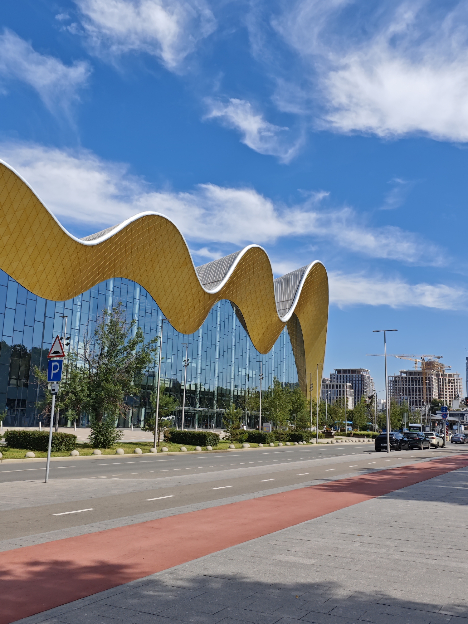 This is a photograph of a modern building with a distinctive wavy, golden roof and a glass facade, located in Moscow, Russia. The building appears to be the Luzhniki Palace of Water Sports, recognizable by its unique architectural design. In the foreground, there is a street with a red bicycle lane and a sidewalk. Several cars are parked or driving on the street. Trees and landscaping are visible between the building and the street. In the background, other buildings, some of which are under construction with cranes, are visible against a blue sky with white clouds. A parking sign is prominently displayed on a pole near the sidewalk. The scene depicts an urban environment on a sunny day.