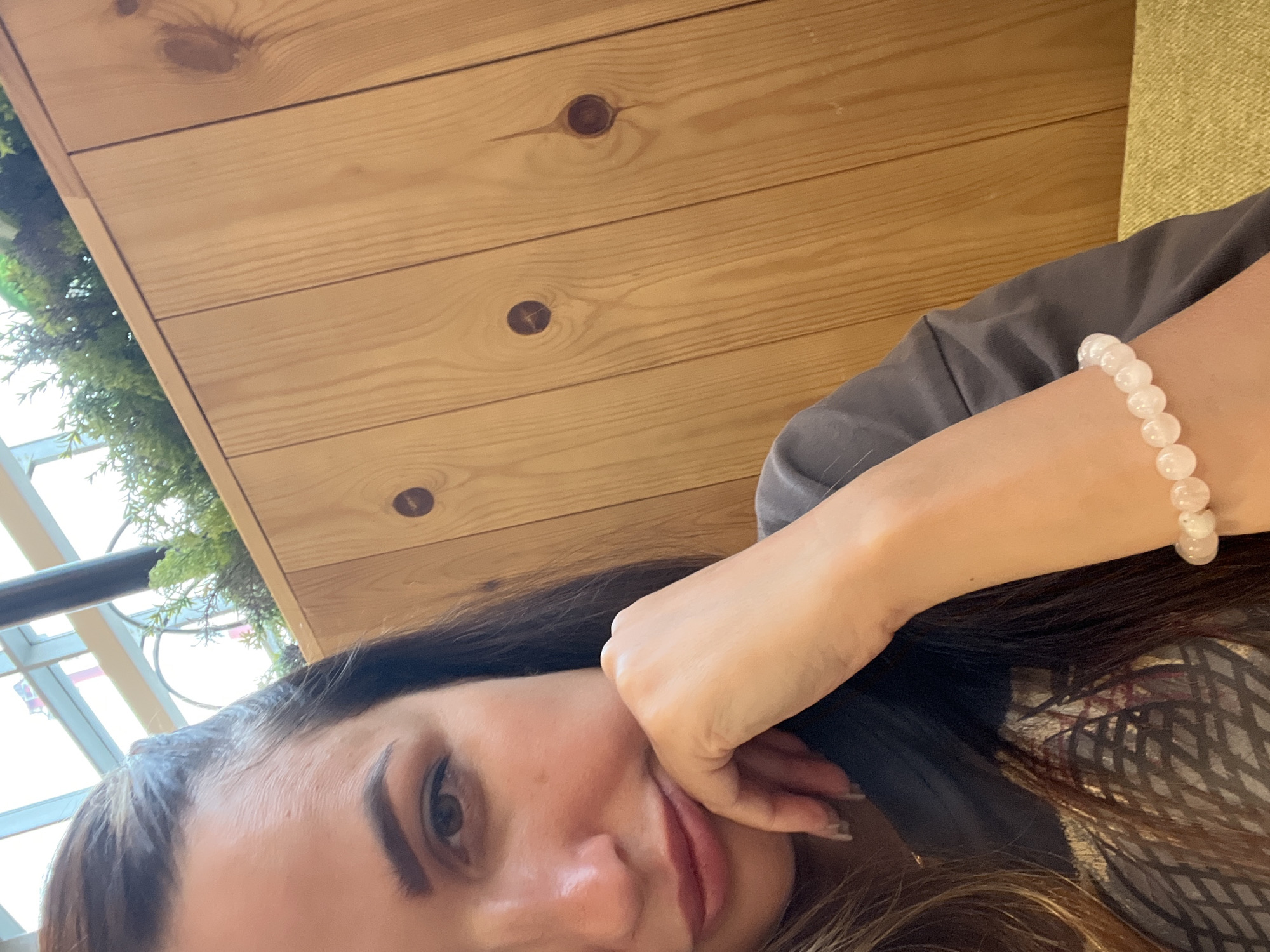 A close-up, waist-up, indoor, eye-level medium shot captures a woman with dark hair, facing the camera and resting her chin on her hand. She wears a dark grey t-shirt and a light pink beaded bracelet on her left wrist. The background consists of a light brown wooden wall with dark circular knots and a blurry green plant in the upper left corner, suggesting an urban setting. The lighting is natural, coming from an unseen source above and to the left.