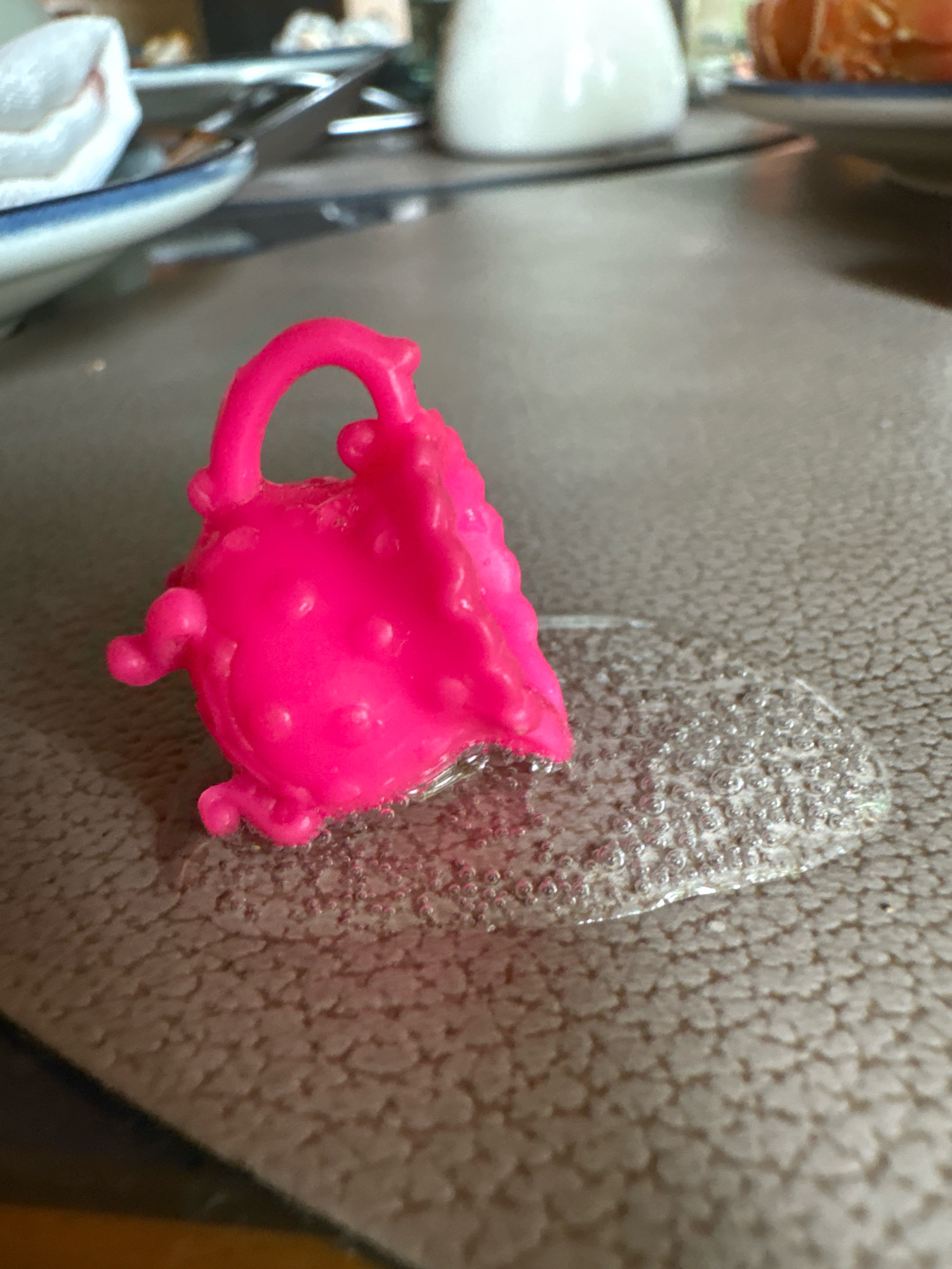 A neon pink, doll-sized purse with a textured exterior and a handle rests on a textured, beige surface. It sits partially submerged in a clear, bubbly liquid. In the background, out of focus, are glimpses of plates with food, a white napkin, and a white pitcher or bottle. The scene appears to be indoors, possibly on a table. The location in Vagator, India, is not discernible from the image.