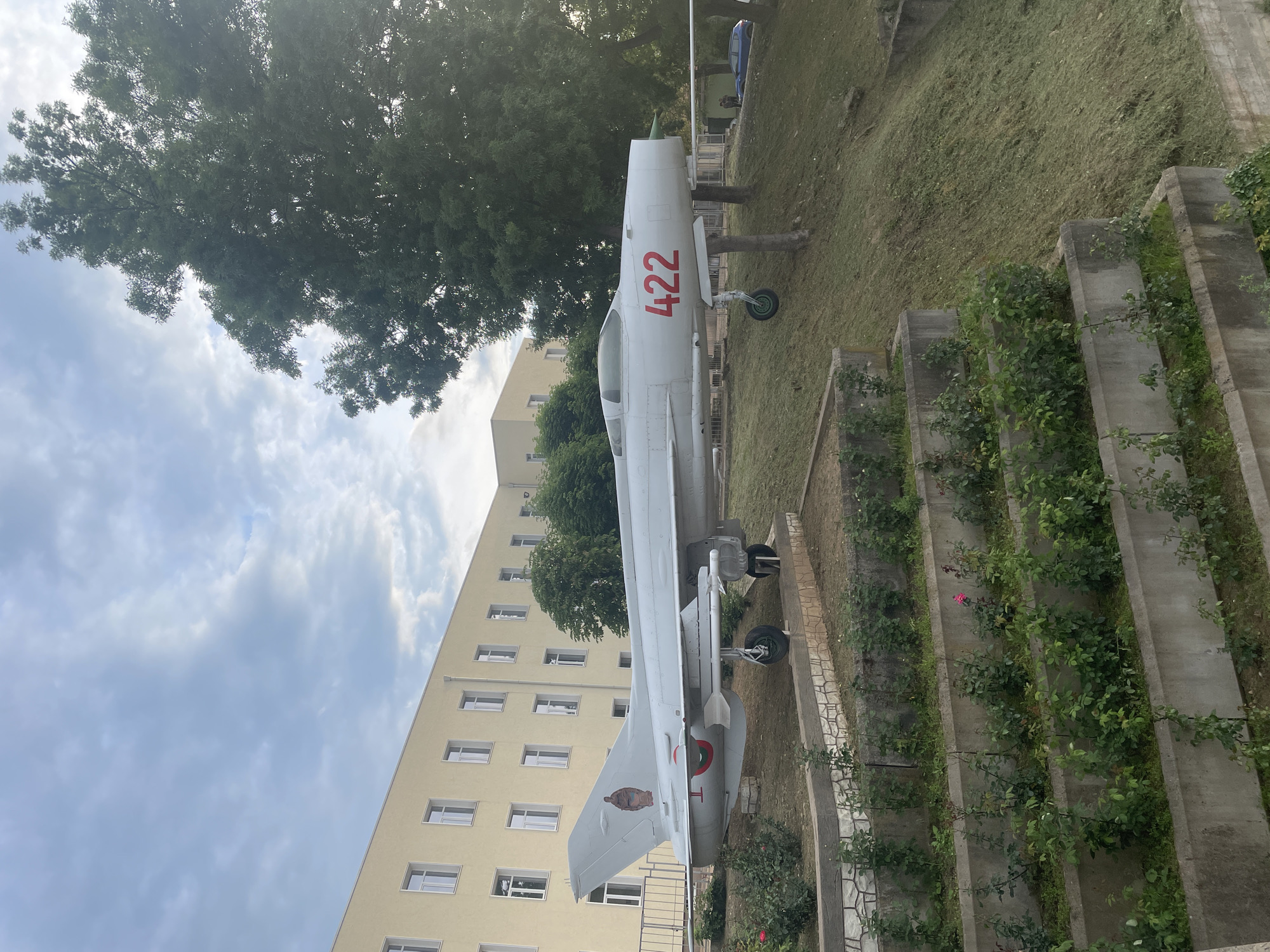 A side view shows an MiG-21 aircraft painted white with the number "422" in red on the fuselage, standing on display at an air park in Unknown, Unknown. The aircraft is parked in a grassy area next to a tiered concrete retaining wall with climbing plants. A light tan-colored building with several windows is in the background. A tall green tree stands behind the aircraft and to the left of the building. The sky is visible above the building and trees, showing some clouds.