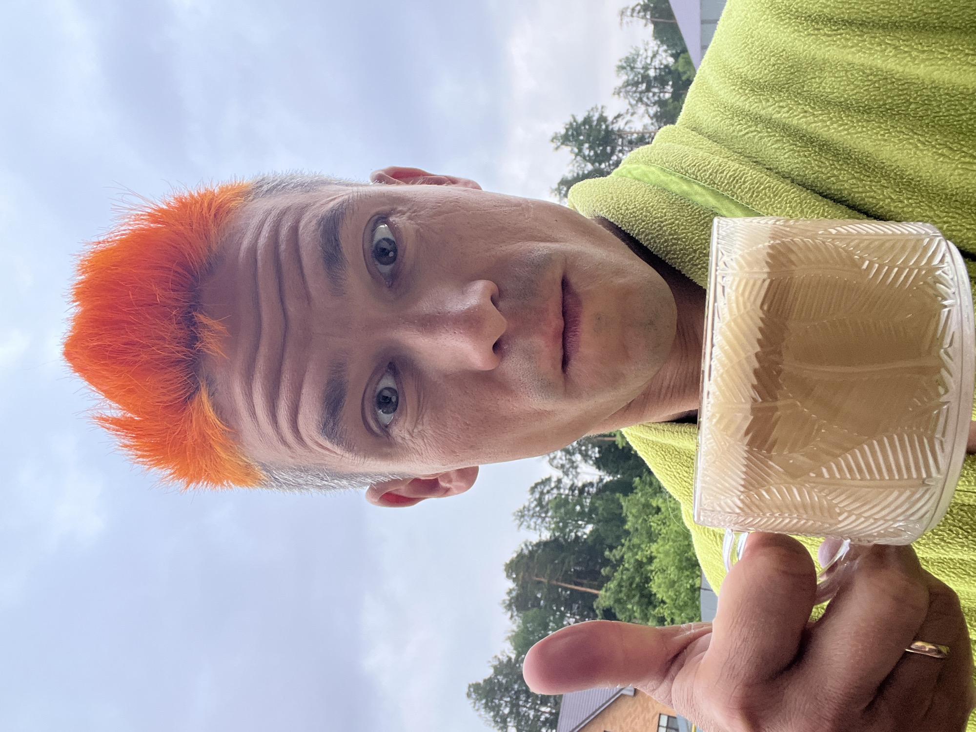 The image shows a fair-skinned person with short, orange hair and blue eyes holding a glass mug filled with a light-colored beverage. The person is wearing a light green fleece jacket. In the background, trees are visible, along with a cloudy sky and the edge of a building with a brown roof. Although the location in the city of Chaikovsky, Russia, is requested, this information is not determinable from the image. There is no information to suggest that this picture was taken in Chaikovsky, Russia.