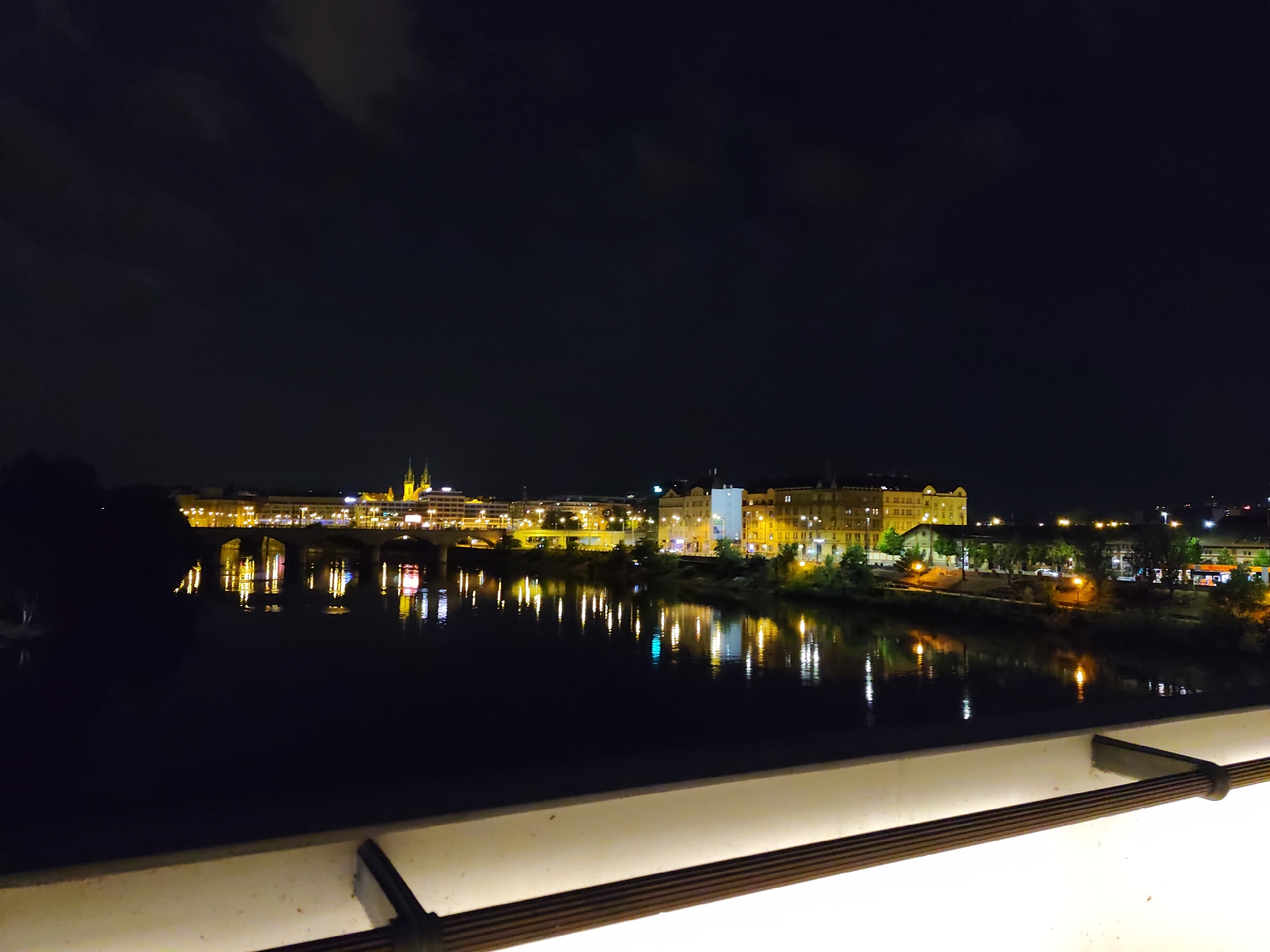 The image captures a nighttime view of the Vltava River in Prague, Czechia, with the illuminated cityscape in the background. A bridge, likely the Mánes Bridge based on its arch structure and proximity to visible buildings, spans the river. The buildings lining the river are brightly lit with yellow and white lights, reflecting on the dark water. Among the illuminated structures, a church with twin spires is prominently visible, suggesting it could be the Church of St. Nicholas on Old Town Square or another significant religious building in the vicinity. The foreground features a white railing with a wooden accent, indicating the vantage point is likely from a balcony, terrace, or a bridge pedestrian walkway. The sky is dark with some diffused clouds, typical of a night scene. No people are discernible in the image. The overall atmosphere is one of urban tranquility and illuminated beauty.