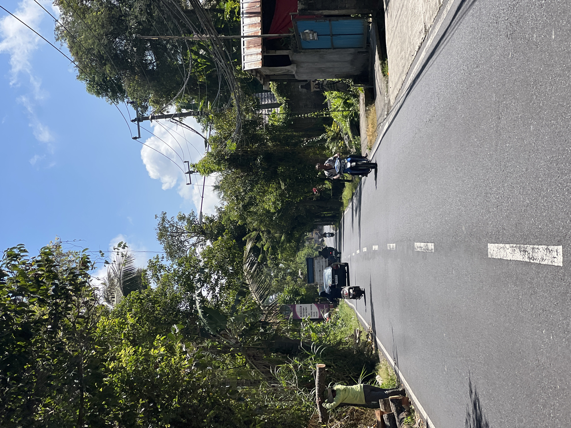 In Payangan, Indonesia, a two-lane asphalt road is lined with lush green trees and vegetation. The sky above is a clear blue with scattered white clouds. On the left side of the road, a person wearing a light green shirt and grey pants carries a large wooden log over their shoulder. Piles of cut logs are visible next to them. A black car and several motorbikes are traveling on the road. One motorbike is being ridden by a person wearing a blue shirt. On the right side of the road, a building with a weathered metal roof and a blue gate is visible, with electrical wires overhead. The overall scene depicts a typical day on a rural Indonesian road.