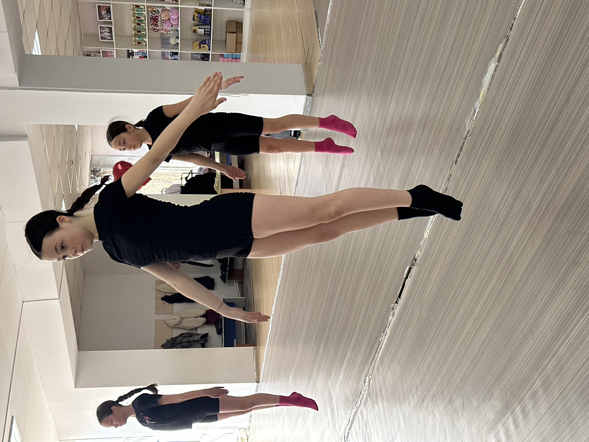 The image shows two young women, one in black socks and one in pink, possibly practicing dance or gymnastics in a studio. Both are wearing black tops and shorts, and their hair is pulled back. They are positioned with their arms outstretched, suggesting a movement or pose. The scene appears to be indoors, indicated by the mirrored wall and ceiling, and the presence of shelving with various items in the background. Based on available information, the location in the city of Votkinsk, Russia, cannot be verified from the image.