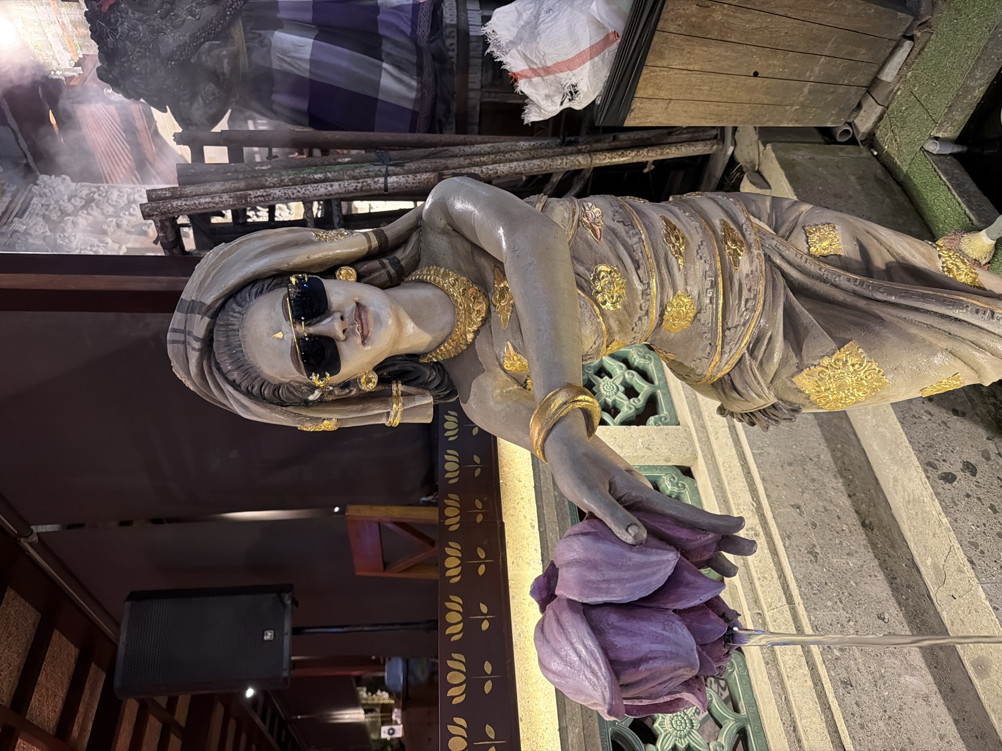 A detailed, full-length female statue, rendered in light beige or taupe with intricate gold leaf patterns, stands prominently in the foreground. Located in the Ubud District, Indonesia, the figure is dressed in traditional-style draped garments, adorned with gold bangles, a gold necklace, circular gold earrings, and a small golden bindi-like embellishment. Her hair is covered by a matching draped headpiece, and her eyes are obscured by modern black wayfarer-style sunglasses with gold frames. She holds a large, multi-petaled purple lotus flower in her extended left hand, from which a thin stream of clear water continuously drips downwards.
The environment suggests a restaurant or cultural space. To the statue's left, a dark wooden railing displays illuminated golden lotus stencils backed by warm lighting. Further left, a black EV speaker on a stand and a red wooden chair are visible. To the right, a cluster of rusty metal poles and scaffolding leans against a wall, with a torn white sack nearby. Behind these elements, a large structure is partly covered by a striped fabric, possibly a Balinese *poleng* cloth, and subtle smoke or haze is present in the upper right. The foreground features concrete steps and a decorative green railing. A small pond with artificial green turf is at the bottom right. A textured, possibly thatched, ceiling with dark wooden beams is visible in the upper left.