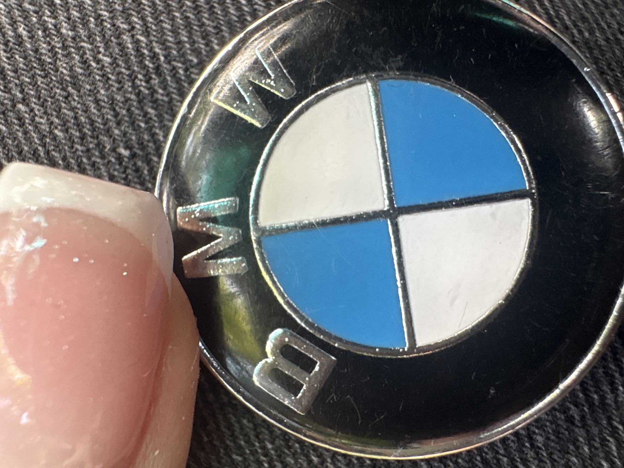 A close-up shot features a human finger interacting with a BMW logo. The finger, positioned on the left, has a French manicure with a pinkish-beige nail bed and a white tip, showing some glitter or shimmer. The BMW emblem is circular, with the iconic blue and white quadrants in the center, surrounded by a black outer ring. The letters "BMW" are visible in silver or metallic text on the black ring, specifically "W" at the top and "B" at the bottom right. The emblem appears to be made of metal and enamel, showing some reflections and minor wear, such as light scratches on its surface. The background consists of a dark, textured fabric, possibly denim or a similar material, providing a contrasting backdrop. The lighting is bright, causing glints on the metallic parts of the logo and the fingernail.