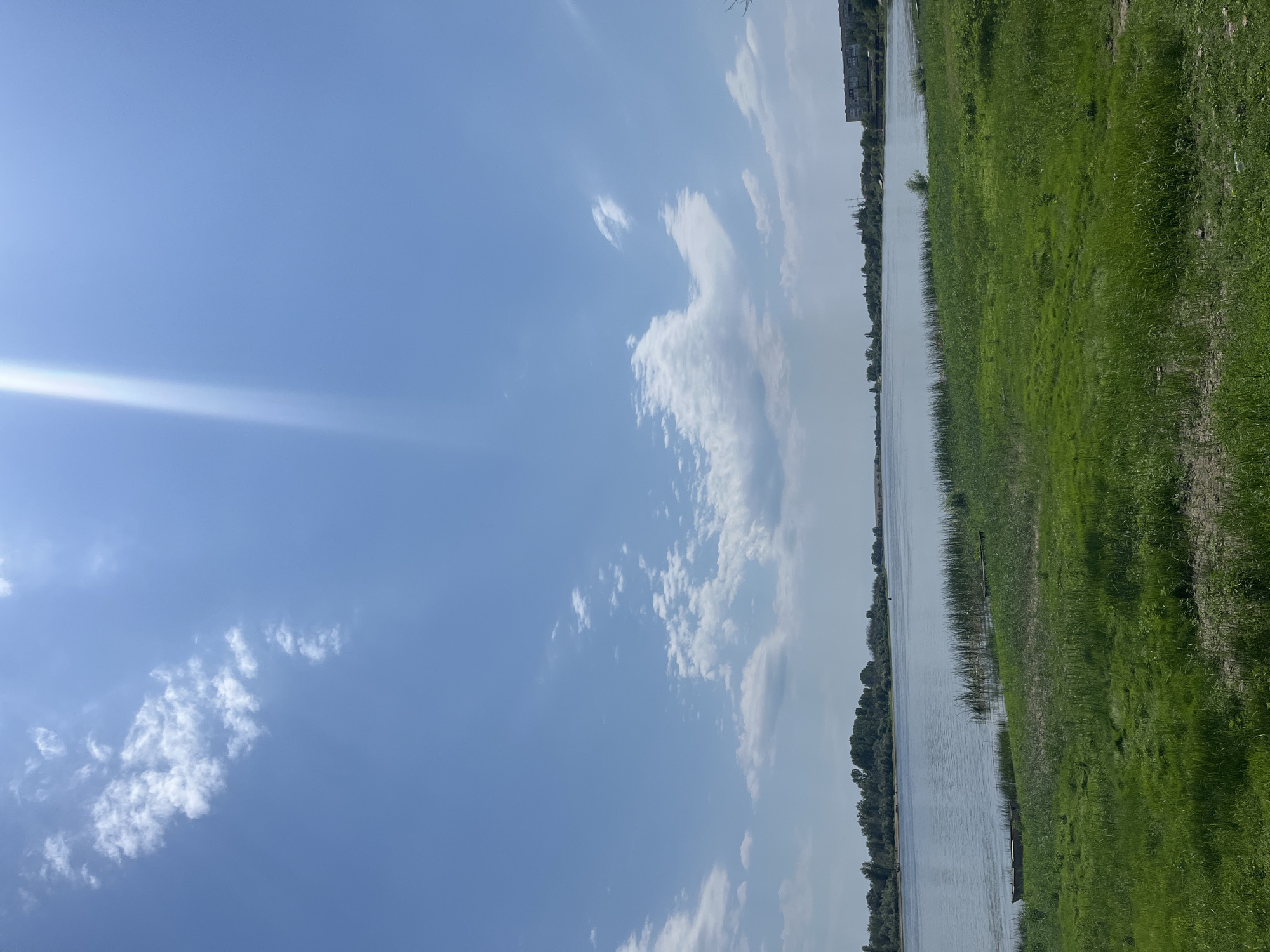 The image depicts a serene, wide river under a bright, partly cloudy blue sky. The sun's rays are visible as a bright streak emanating from the upper left quadrant. The foreground consists of lush green grass with some patches of dry earth. Along the riverbank, tall reeds and grass grow, with two small, dark, empty boats partially submerged near the shore. Across the river, a densely wooded treeline stretches horizontally, with a distant urban settlement visible on the right bank, characterized by low-rise buildings and some industrial structures. The overall impression is a tranquil natural landscape with signs of human habitation in the distance. The scene is consistent with a location in Krasnyye Barrikady, Russia, likely near the Volga River delta. There are no people or animals visible in the image, and no actions or interactions are taking place.