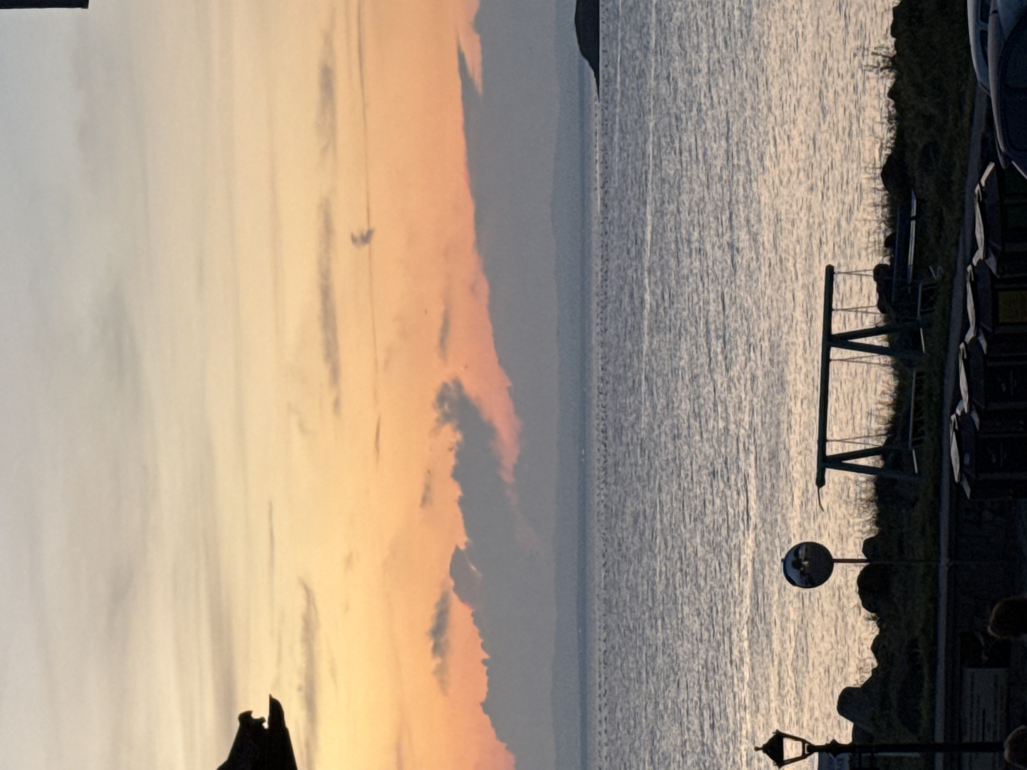 A silhouette of a gargoyle-like structure, possibly a decorative element on a building, is visible on the left side of the frame against a pastel sunset sky. The sky transitions from pale orange near the horizon to a soft blue at the top, with scattered wisps of clouds. In the middle ground, a calm body of water, likely the Black Sea, stretches across the image, with a faint line of buoys or markers visible on its surface. In the distance, a hazy landmass or mountain range can be seen along the horizon, and a small island is situated to the far right.
The foreground depicts a shoreline with dark, rough-textured rocks and sparse vegetation. A street lamp with an ornate design stands on the left. A circular convex mirror on a pole is positioned near the center. To the right of the mirror, a swing set with two seats is present, and a bench is partially visible behind it. Further to the right, several dark, rectangular objects, possibly trash bins or storage units, are lined up. The bottom edge of the frame shows the dark silhouettes of people and the tops of parked cars. The overall scene suggests a coastal area at dusk, with man-made elements like the swing set and street lamp contrasting with the natural landscape. The location is not specified to be Sozopol, Bulgaria in the image.