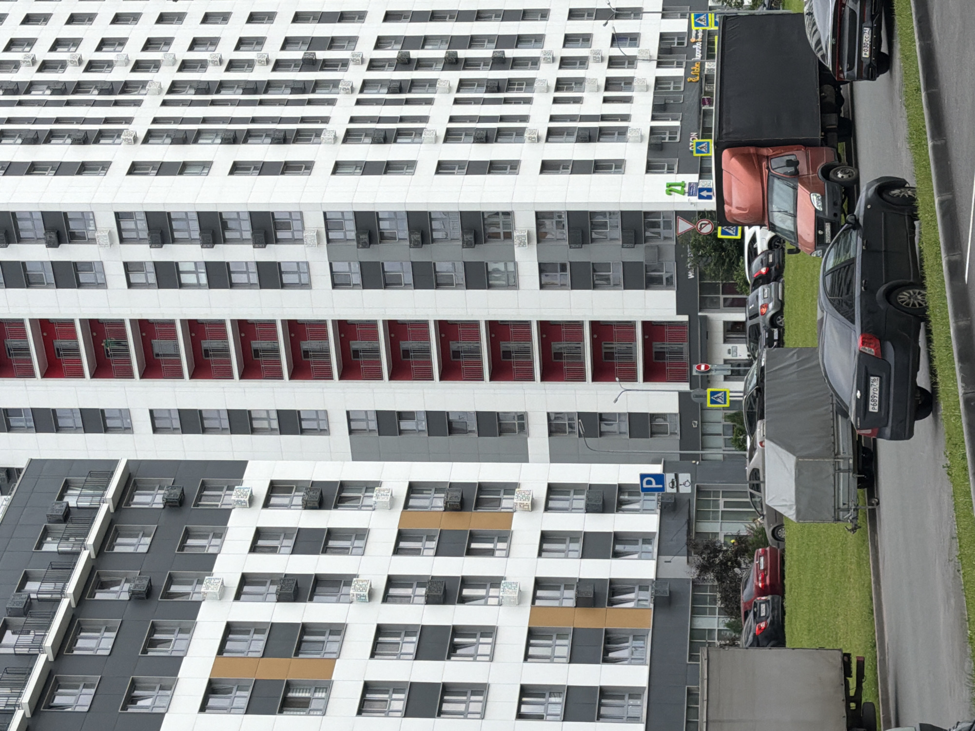 The image depicts a street scene in Kazan, Russia, featuring a modern, multi-story apartment building with a facade of white, gray, and red sections. The building has numerous windows and balconies. In front of the building, several vehicles are parked along the roadside, including a red truck, a gray trailer, a black car, and other cars partially visible. The scene appears to be a residential area.
The time of day is likely daytime, with overcast or diffused lighting, suggesting it could be a cloudy day. There are no people visible in the frame. The primary activity is parking, with vehicles lined up next to the building and along the road.
Notable details include a blue parking sign with a "P" symbol, a yellow road sign with a warning symbol (likely a pedestrian crossing), and another road sign with a red circle and a diagonal line, indicating a prohibition. The license plate of the black car is partially visible as "P689YO716". The building's architecture is contemporary, with repetitive window patterns and the use of contrasting colors. The presence of air conditioning units on the exterior of the building is also a detail. The ground is covered with green grass and asphalt, indicating an urban or suburban setting.