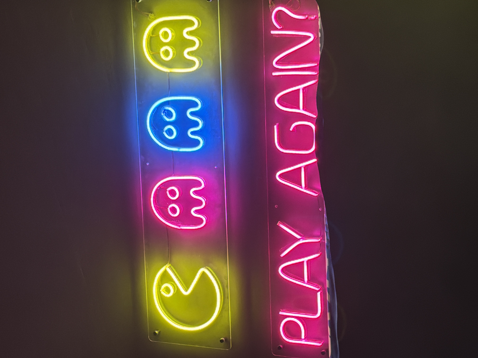 The image shows two vertical neon signs against a dark background. The left sign displays a sequence of four Pac-Man characters/ghosts in different colors: yellow, blue, pink, and yellow (from top to bottom). The right sign spells out "PLAY AGAIN?" in pink neon letters. The signs are affixed to the wall with visible screws. There is no indication of Izhevsk, Russia in the image.