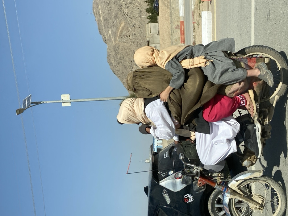 The scene depicts three individuals riding an older model motorcycle on an asphalt road in Kandahar, Afghanistan, under a clear blue sky.
The lead individual, a male, is operating the motorcycle. He wears a white long-sleeved shirt, white traditional trousers (salwar), a dark vest, and a light beige headscarf covering his hair and neck. His footwear consists of dark athletic shoes.
Seated directly behind the driver is a second individual, an adult female, largely obscured by a dark olive-green burqa or chador. Visible beneath the garment are red traditional trousers (salwar) with a white embroidered pattern near the ankle, and light brown flat sandals.
A third individual, a child, is seated at the rear of the motorcycle. The child wears a grey traditional long tunic and trousers, a light yellow headscarf, and grey slip-on shoes. The child's left arm is wrapped around the torso of the middle passenger.
The motorcycle has a red frame and a chrome front fender. The road surface is asphalt, with a concrete barrier visible on the right side of the frame. In the left background, a black car with stickers, including "CTC" and a heart-eyed skull, is partially visible. Further in the background, an aluminum pole with a solar panel and a street light fixture stands under power lines. Arid, rocky mountains with a winding path are visible in the distance on the right side of the frame. Several indistinct buildings are also present in the middle ground.