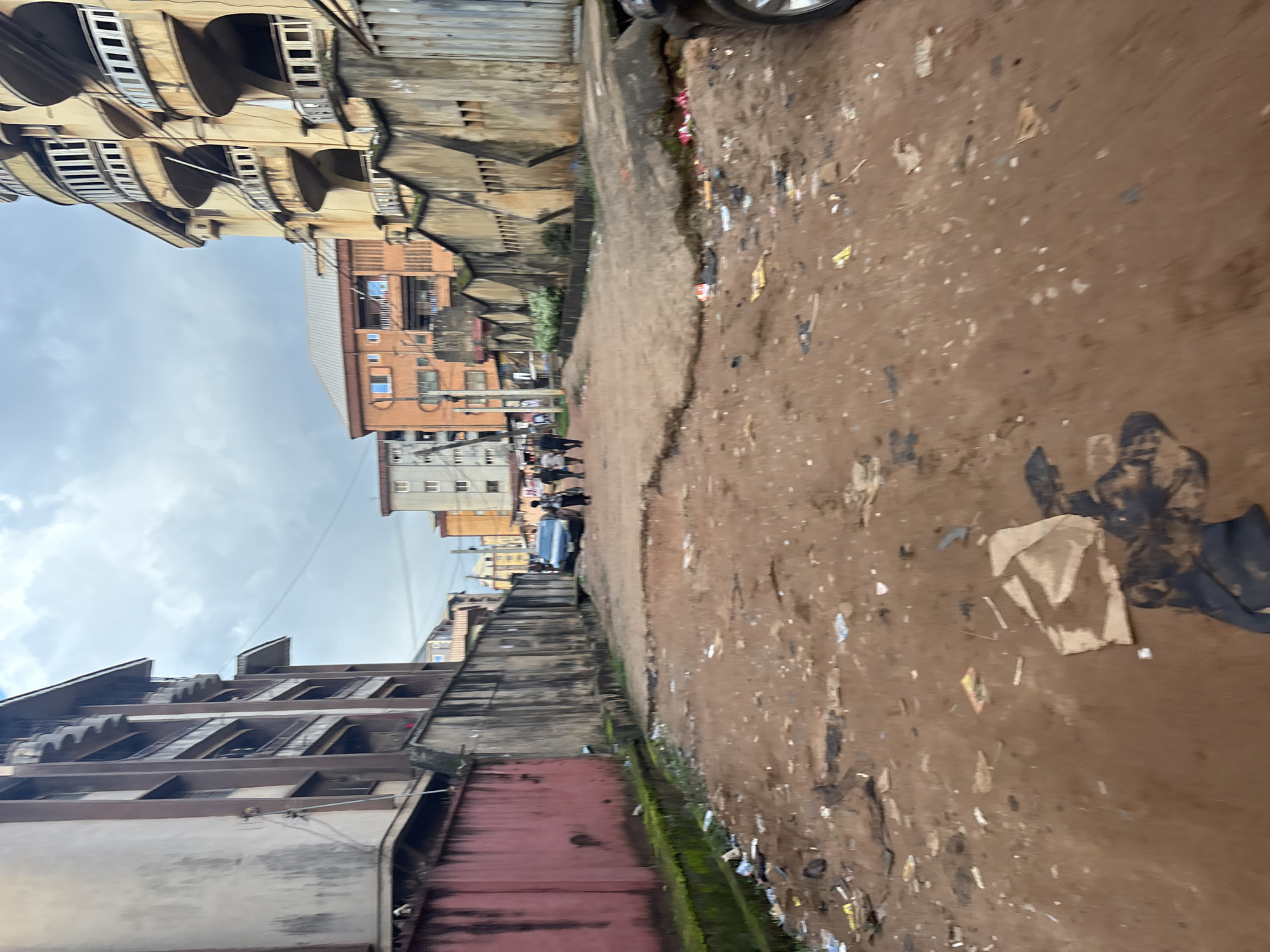 A ground-level, eye-level shot captures a scene of urban decay in Onitsha, Nigeria. The foreground is dominated by a dirt and debris-strewn path, littered with discarded paper, plastic, and organic waste. A large piece of torn cardboard lies prominently in the center foreground, with dark, amorphous masses of refuse scattered around it. To the left, a weathered, red-painted concrete wall extends into the background, with moss growing along its base and a narrow, debris-filled trench.
In the midground, a dark sedan is parked on the right side of the path, partially obscuring a concrete barrier with a textured, zigzag pattern. Several people, appearing to be adults, are walking away from the camera towards the left midground, silhouetted against the buildings.
The background features a street lined with multi-story buildings. On the left, a light-colored building with visible balconies and windows is partially obscured by a darker, textured wall. Further back, an orange building with a flat roof and numerous windows stands out, with other structures of varying colors and architectural styles visible behind it. The sky above is overcast with heavy, gray clouds.