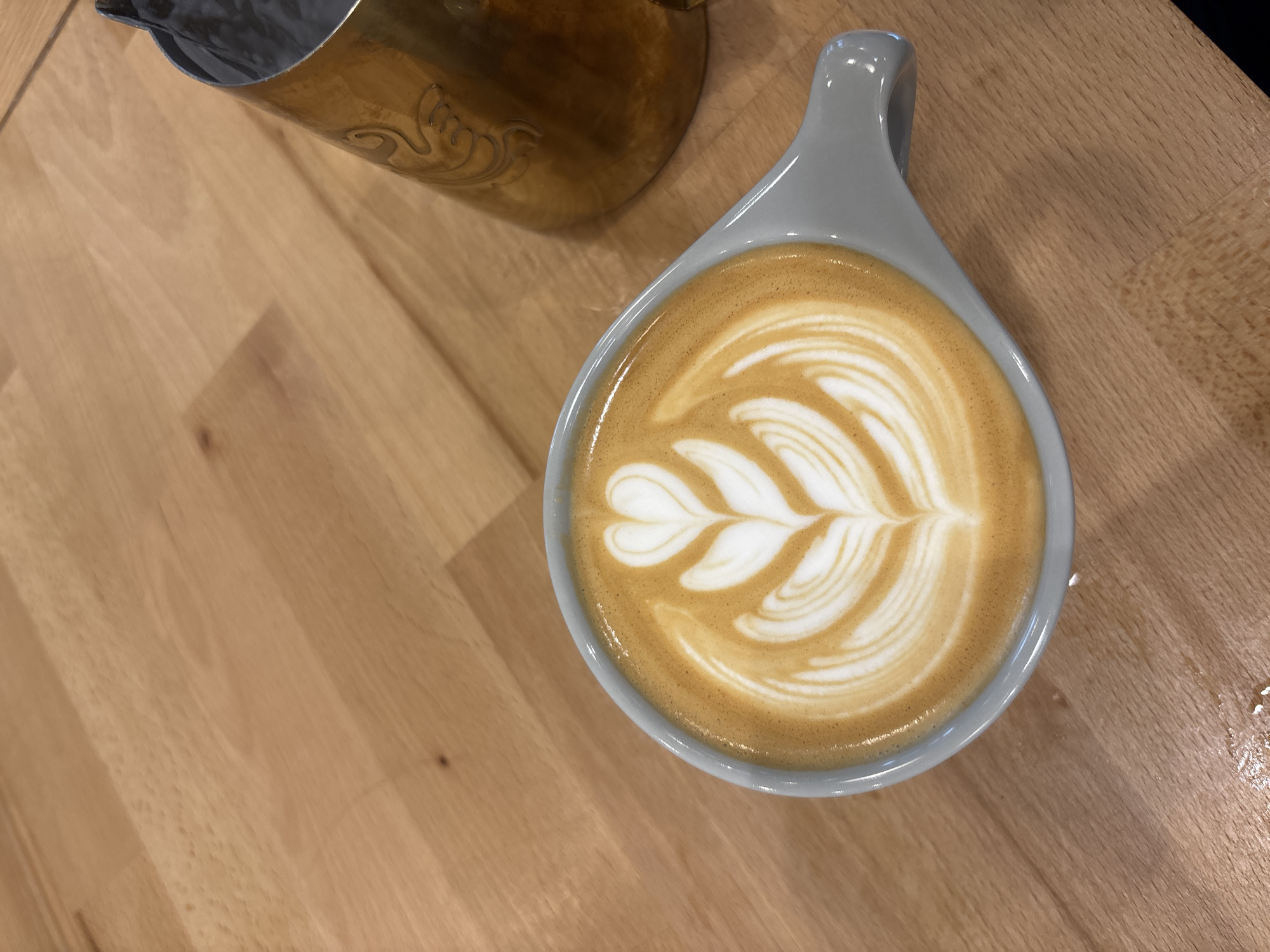 An overhead shot captures a beautifully crafted coffee beverage, likely a latte or cappuccino, resting on a light-colored wooden surface. The coffee is presented in a modern, light grey ceramic cup with an elongated, integrated handle. Dominating the coffee's surface is an intricate latte art pattern, a classic "rosetta" or "fern" design, culminating in a delicate heart shape at its top. The white foam stands out against the rich, warm brown of the coffee, indicating a freshly poured drink and skilled barista work.
Partially visible in the upper left corner is a metallic pitcher, possibly brass or bronze, featuring an elegant, etched decorative motif on its side. This object suggests the immediate context of coffee preparation, likely a milk frothing pitcher. The wooden surface beneath both items appears to be a clean countertop or table with visible grain, indicative of an indoor setting.
The scene conveys a calm atmosphere, focusing on the aesthetic appeal of the coffee. There are no people visible, nor is there any discernible text. The lighting is even, highlighting the textures and colors of the objects. This tranquil moment takes place in Sofia, Bulgaria.