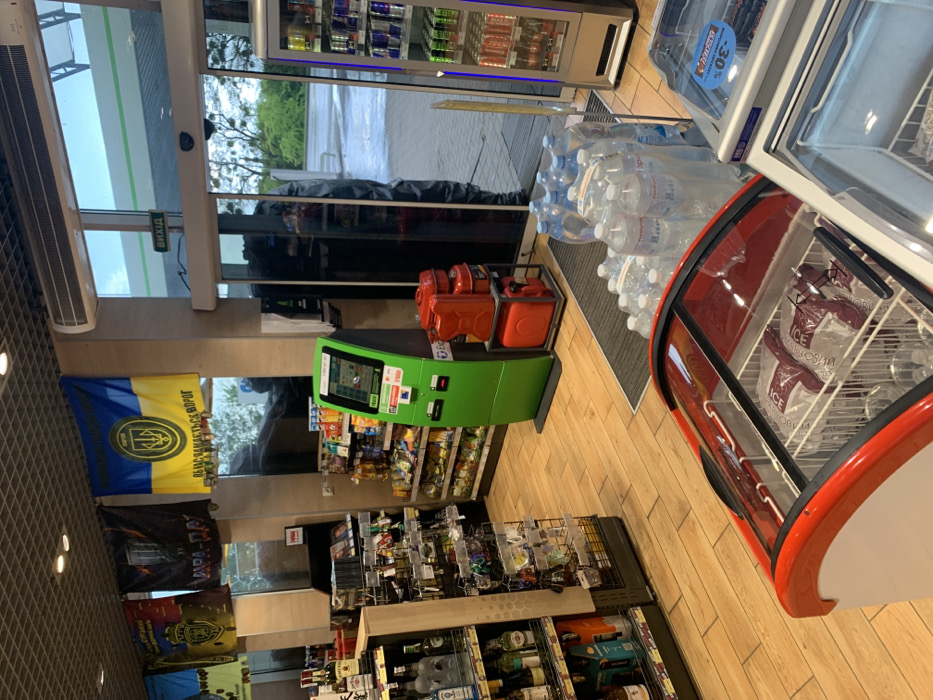 The image shows the interior of a convenience store in Solonytsivka, Ukraine (latitude: 49.973, longitude: 36.02). The store is relatively small, with wooden floors, and a variety of goods on display. Two glass-front refrigerators sit in the foreground, one containing packages of frozen food, the other empty. A large green ATM machine is visible in the center of the image. To the left, a wall of shelves displays a variety of alcoholic beverages, including bottles of vodka, wine, and beer. A Ukrainian flag hangs on the wall behind the ATM machine. A glass door marked "вихід," meaning "exit" in Ukrainian, is visible in the background, allowing a view outside the store. The entrance to the store is just beyond the ATM machine, and the door to the right of the ATM machine is open. A person can be seen standing behind the counter. The store appears to be well-lit, and the temperature appears to be moderate.