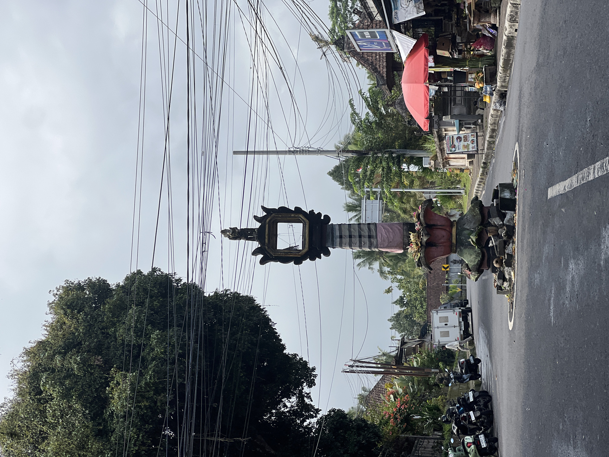 This image depicts a street scene in Tembuku, Indonesia, likely at an intersection or roundabout. Dominating the center is a tall, ornate monument, possibly a religious or cultural marker, with intricate carvings and draped fabric. It's surrounded by loose stones and offerings at its base. To the left, a large, leafy tree shades a collection of parked motorcycles. Beyond the tree, a white truck is visible, and further on, traditional Balinese-style buildings with tiled roofs line the street. On the right side of the road, a red umbrella shelters a small market stall displaying fruits and goods, with various shops and structures behind it. The street itself is asphalted, with white lane markings visible. Overhead, a dense network of power lines crisscrosses the sky against a cloudy, overcast backdrop. A few individuals are subtly present; one person walks near the white truck, and others are likely present within the market stalls, though not clearly defined. The environment appears to be a somewhat humid and tropical locale.