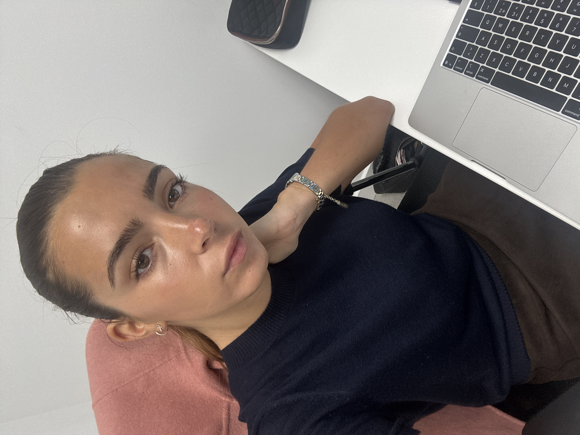 A medium-shot, angled selfie captures a woman seated at a white desk with a laptop. She has dark hair pulled back, fair skin, and wears a dark blue sweater. A pink garment is visible beneath the sweater on the left side. A silver bracelet adorns her wrist. The laptop, positioned on the right side of the frame, features a visible keyboard and trackpad. The desk also holds a small black case in the upper left corner. No details indicate a specific location in Torres Vedras, Portugal. The setting appears to be indoors.
