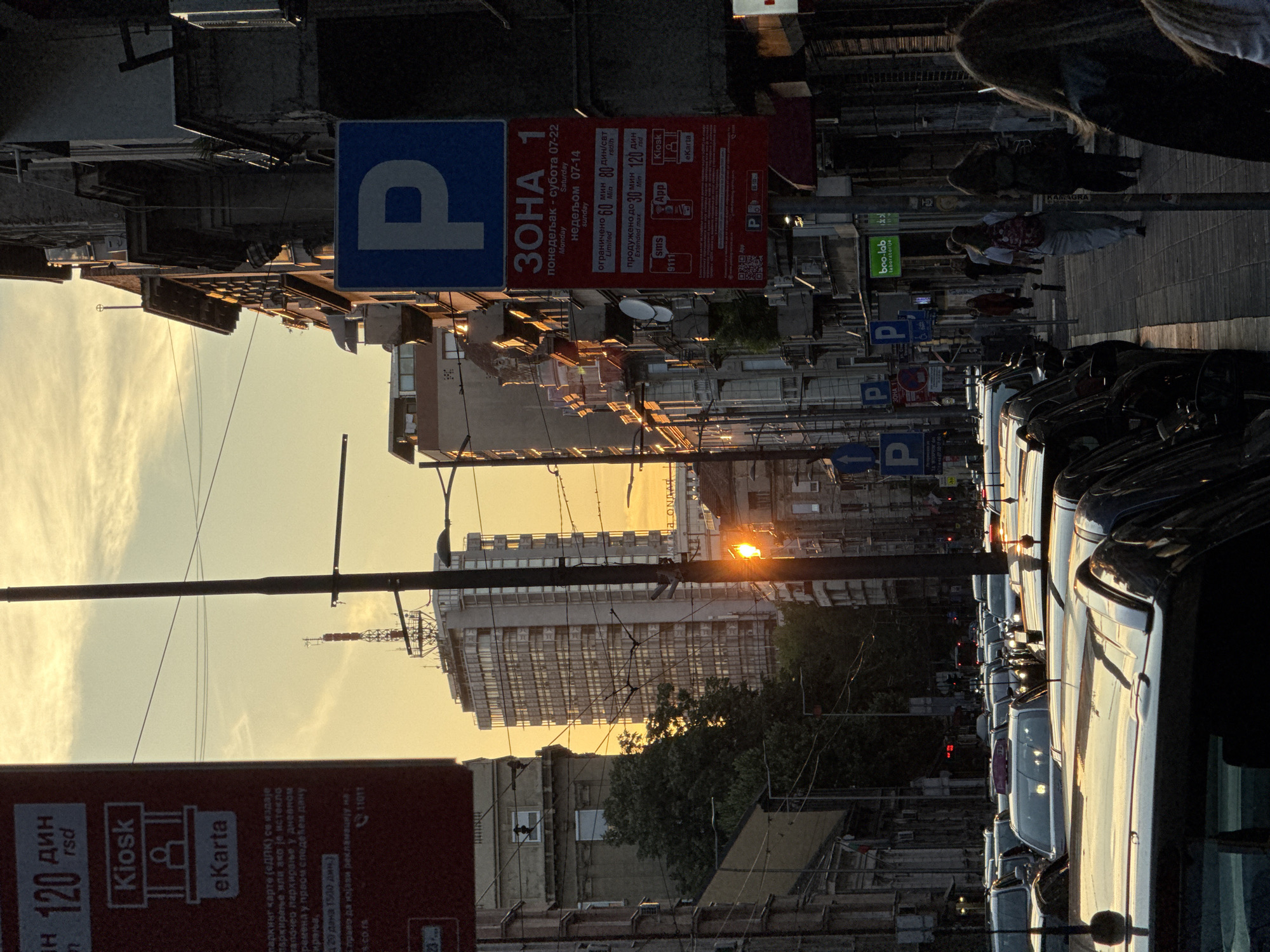 The image depicts a street scene in Palilula Urban Municipality, Serbia, during sunset. The low-angle sunlight casts a warm glow on the buildings and reflects off the wet asphalt, indicating recent rain. A line of parked cars, mostly black, occupy the foreground and extend down the street, with some showing reflections of the sky. A tall, modern building with a glass facade stands prominently in the background, topped by a communication tower. Another older, lighter-colored building is situated to its left.
Several street signs are visible, including a blue parking sign with a white "P" and a red sign indicating "ЗONA 1" (Zone 1) with parking regulations in Serbian. Some of these signs have Cyrillic text, suggesting a location in Serbia. A kiosk advertisement is also partially visible on the left. A few pedestrians are walking on the sidewalk on the right side of the street, some carrying backpacks. Overhead, a complex network of electrical wires crisscrosses the sky, typical of urban infrastructure. The overall atmosphere is one of a bustling city street at dusk, with the warm light creating a dramatic and slightly hazy effect.