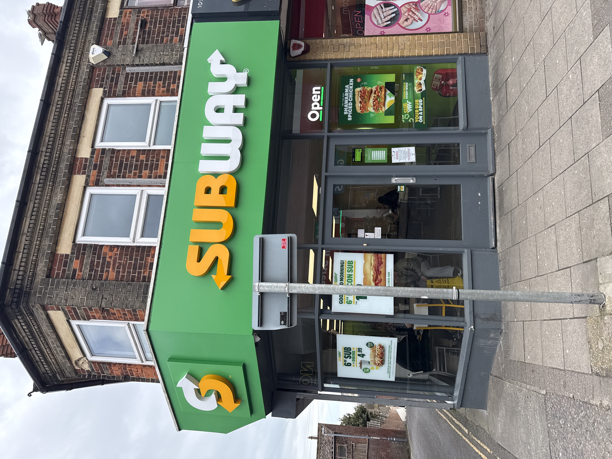 The image shows the exterior of a Subway restaurant located at 109 High Street, Gorleston-on-Sea, United Kingdom. The building is a two-story brick structure with white-framed windows. Above the gray-framed entrance, there's a green awning with the Subway logo in white and orange. A pole and sign are visible in front of the entrance, partially obscuring promotional posters in the windows. The posters advertise 6-inch sub deals and a shawarma spiced chicken sub. An "Open" sign is visible in the window. The sidewalk in front of the store is paved with gray concrete blocks. There is another store (nails) next door. A person is sitting in the interior.