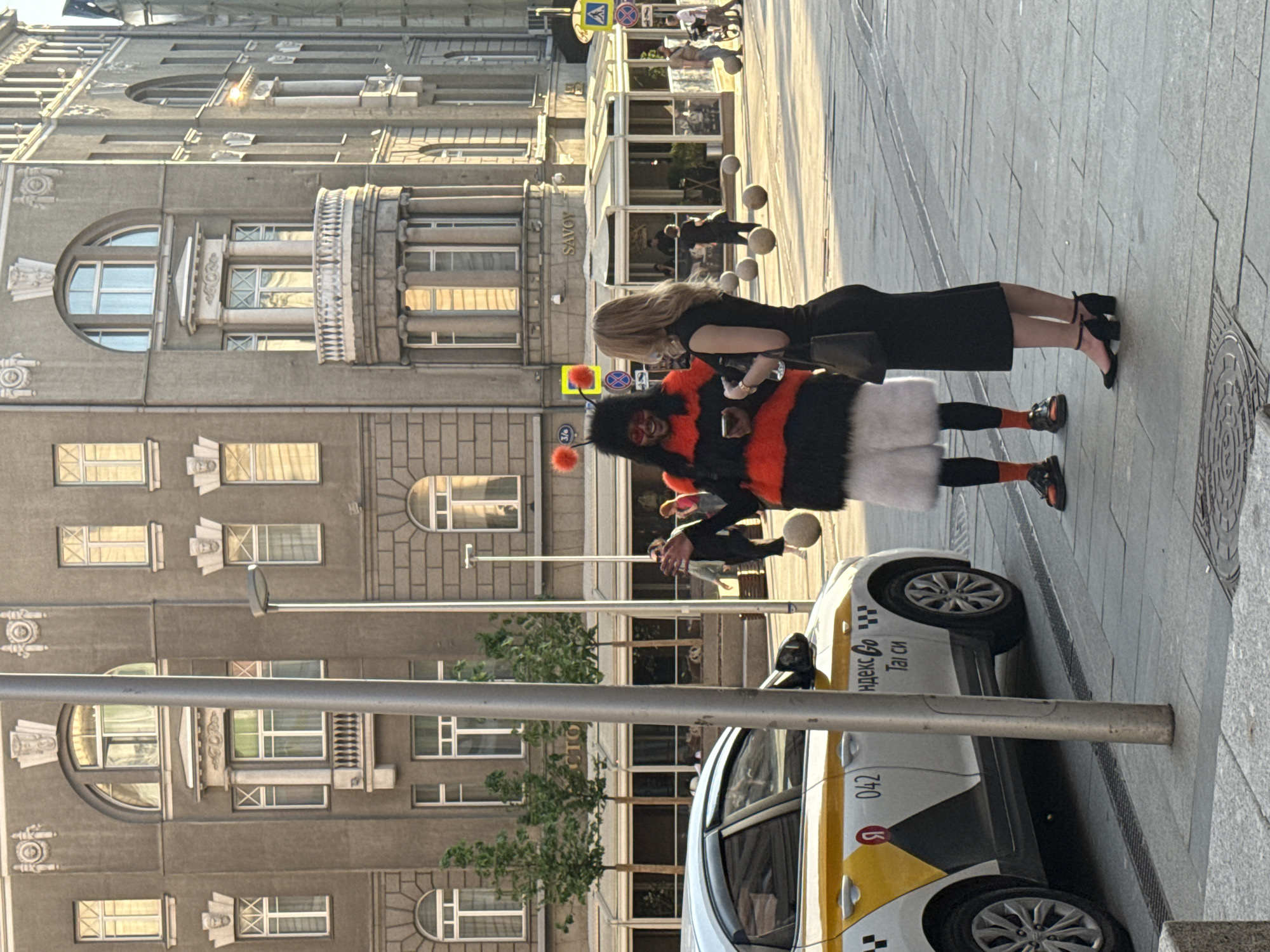The scene is a street in Moscow, Russia. A person dressed in a large black and orange bee costume with furry white "legs" is the focal point, standing on the sidewalk. They are next to a taxi cab which has white and yellow paint and text in Russian. The person in the costume is interacting with a woman in a black dress. The architecture of the buildings in the background appears to be historic. Signage for "Savoy" can be seen on the building.