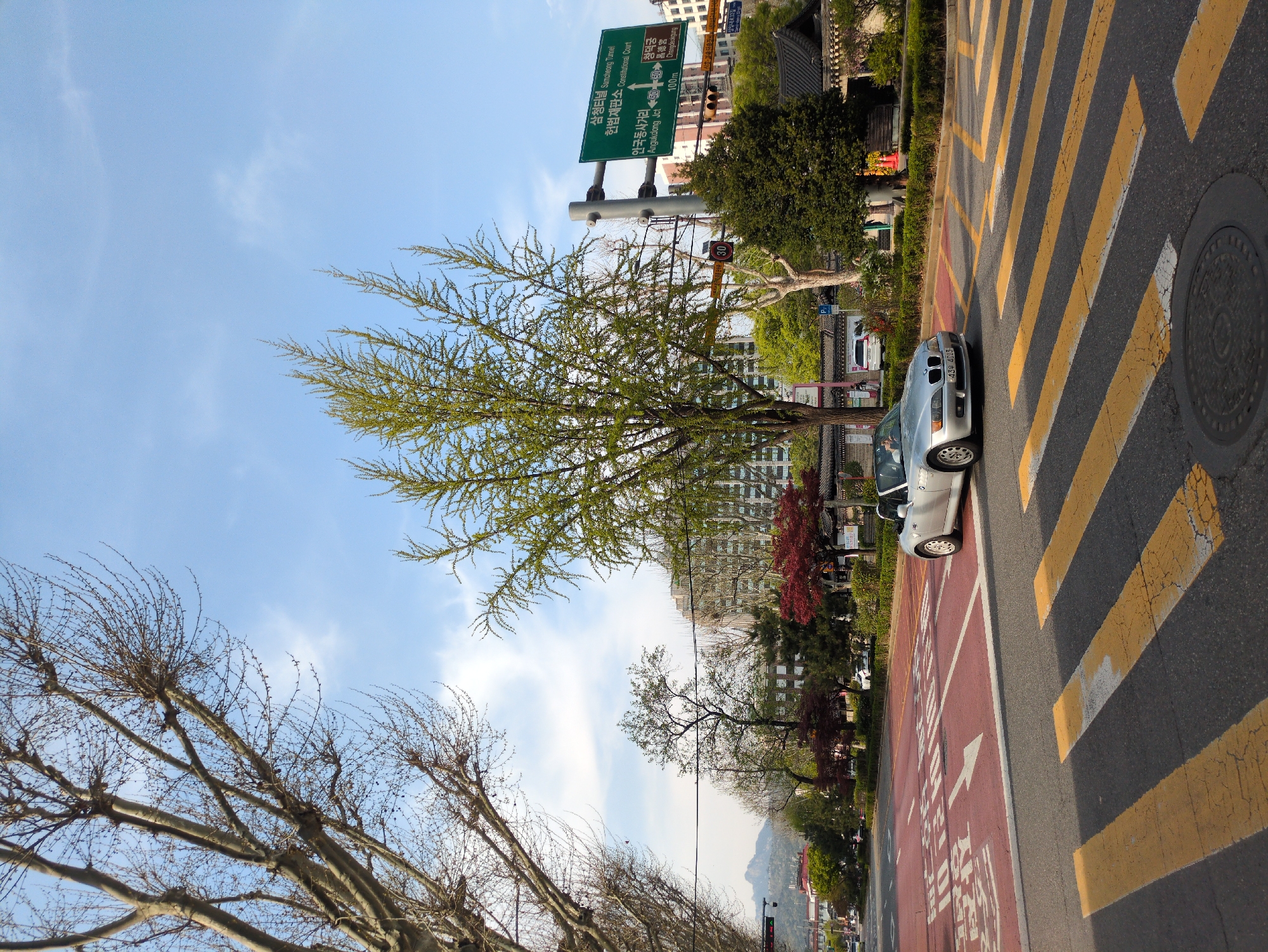 The image depicts a street scene in Seoul, South Korea. A silver car is parked along the roadside, next to a red lane marked with an arrow. The road has yellow zebra stripes. A tree with sparse green leaves stands to the left. A road sign overhead points to "삼청터널 Samcheong Tunnel" and "헌법재판소 Constitutional Court" on the left and "창덕궁 昌德宮 Changdeokgung" on the right. In the background, there are buildings, trees, and a mountain range visible. The sky is blue with scattered clouds.