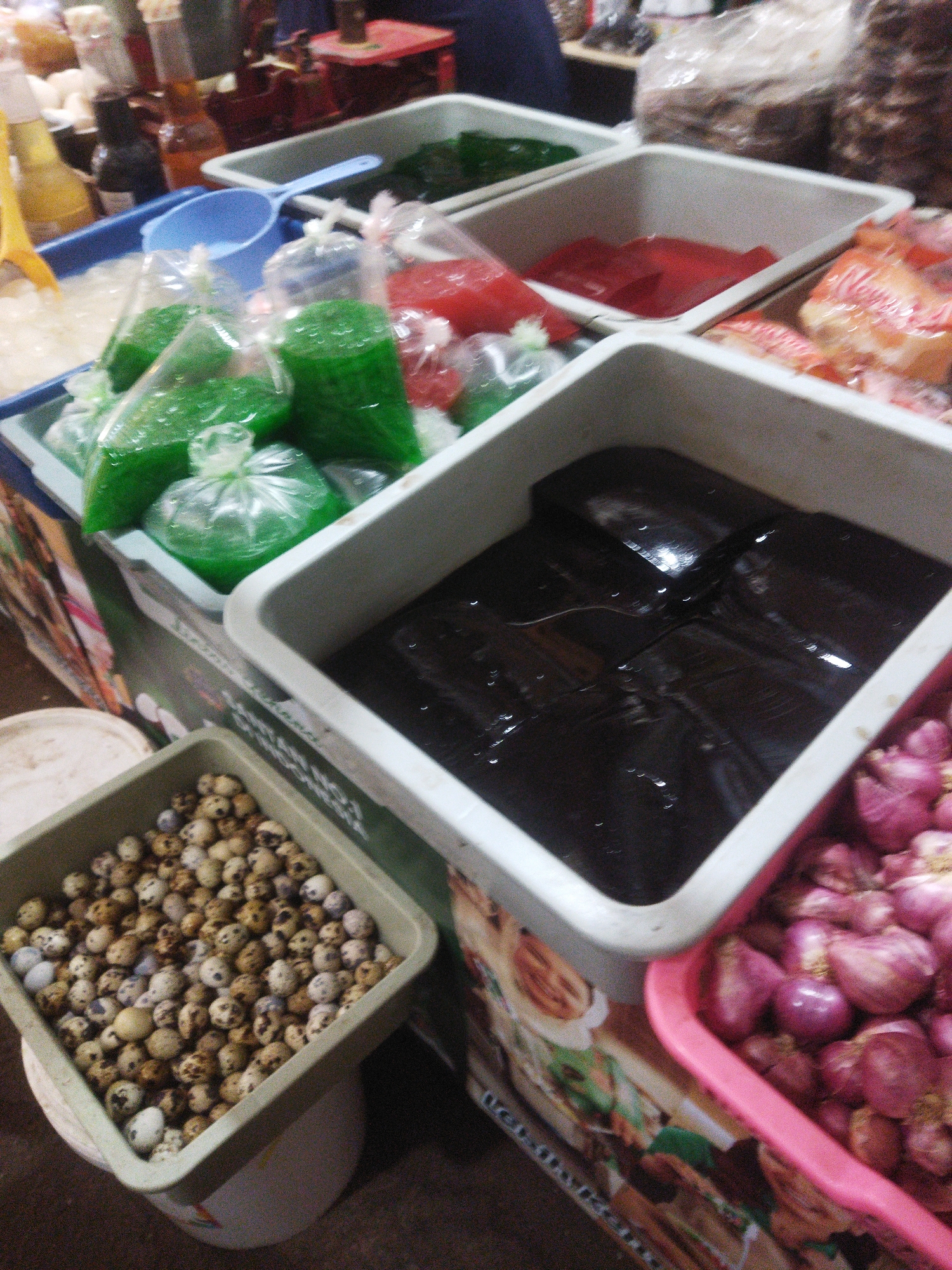 The image depicts a market stall in Wonogiri, Indonesia, showcasing various food items. In the foreground, a green container holds numerous small, speckled quail eggs. To its right, a pink container is filled with red onions. Behind these, a large white container holds a dark, gelatinous substance, possibly a dessert or sweet treat. Further back, to the left, is a blue basin with a blue scoop, containing ice and possibly some liquid. Several plastic bags filled with bright green, translucent jelly are visible in this basin. Next to this, another white container holds red jelly cubes. In the background, shelves are stacked with bags of dry goods, and bottles of condiments are also visible. The overall impression is that of a bustling local market, with an emphasis on fresh produce and traditional Indonesian sweets.