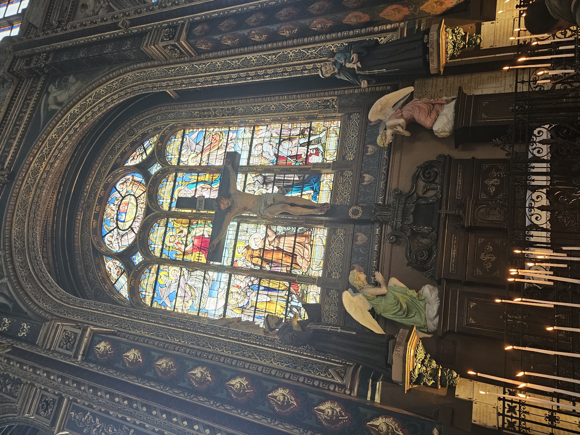 The image shows the interior of Sainte-Chapelle in Paris, France. The photograph is taken at an angle, highlighting the ornate details of the architecture. The scene centers on a large stained glass window depicting religious scenes, above which a crucifix hangs. Flanking the window are sculptures of angels and saints. Rows of lit candles are visible on the right side of the frame. The walls and ceiling are intricately decorated with carvings and paintings.