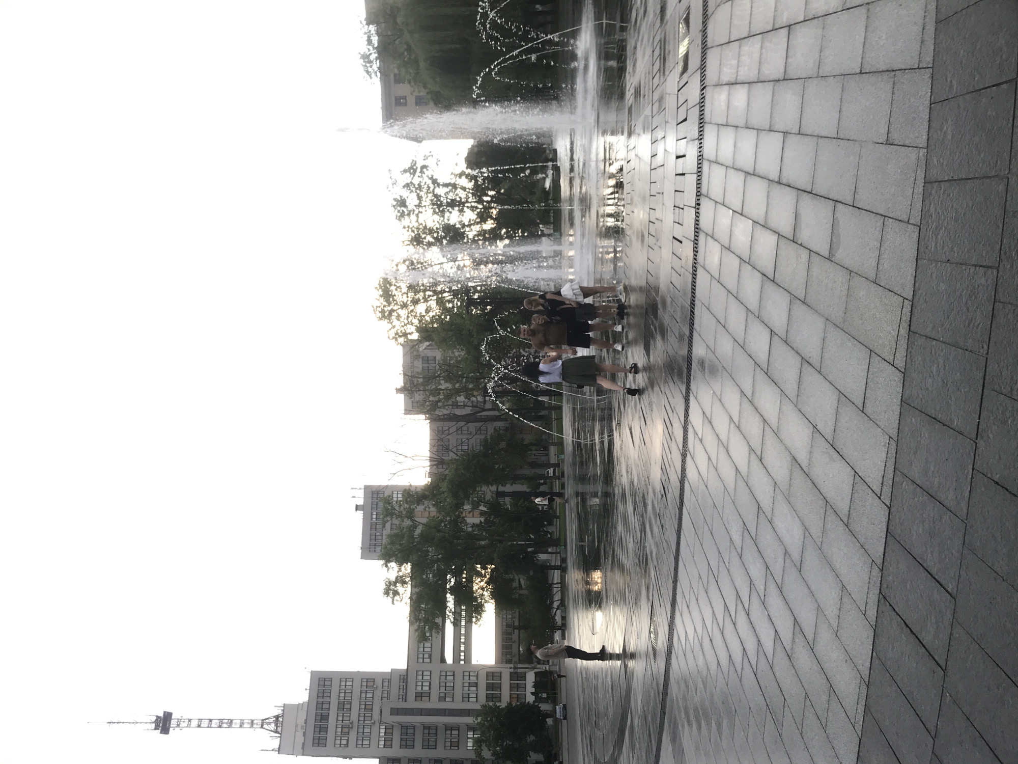 Here's a concise description of the image:
The photograph captures a water fountain display in Kharkiv, Ukraine. Several people are near the fountain, with a group standing directly within the water jets. The ground is paved with light gray stone tiles and appears wet, reflecting light. Buildings, including a tall structure with an antenna, are visible in the background amidst trees. The sky is bright, possibly indicating daylight.