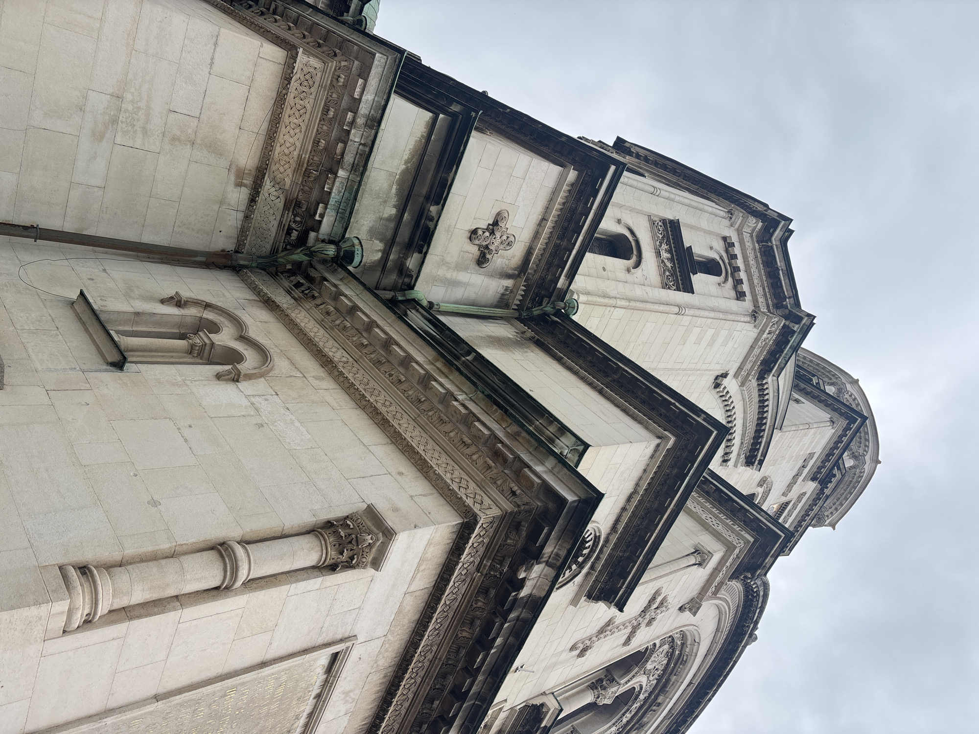 The image shows the exterior of the St. Alexander Nevsky Cathedral in Sofia, Bulgaria, taken from a low-angle perspective. The architectural details of the facade are visible, including stone blocks, decorative carvings, windows, and cornices. The sky is overcast. No people are visible in the frame.