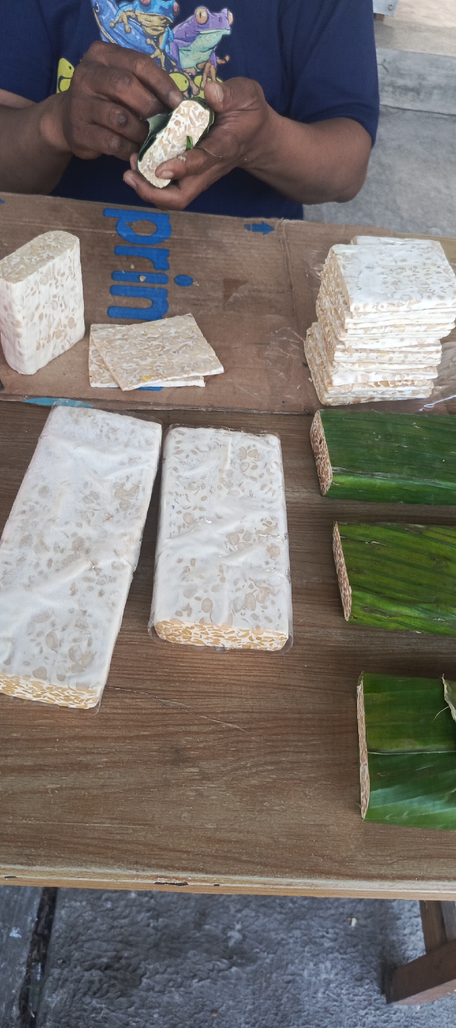 In Sukaresmi, Indonesia, a person wearing a blue t-shirt with a frog graphic is in the process of preparing tempeh. The tempeh is a fermented soybean product, and the individual is shown holding a piece, seemingly inspecting or manipulating it.
The scene takes place outdoors, likely at a market or roadside stall, indicated by the rustic wooden table and the presence of what appears to be concrete flooring in the background. Various forms of tempeh are displayed on the table. There is a single block of tempeh on the left, and a stack of smaller, square pieces of tempeh to the right. In the foreground, two larger blocks of tempeh are individually wrapped in clear plastic. To the right of these, several portions of tempeh are wrapped in green leaves, possibly banana leaves, which is a traditional method of packaging tempeh. The background includes a brown cardboard surface with blue markings, some of which resemble letters and arrows. The overall impression is that of a food preparation or sales environment, with an emphasis on the fresh, handmade quality of the tempeh.
