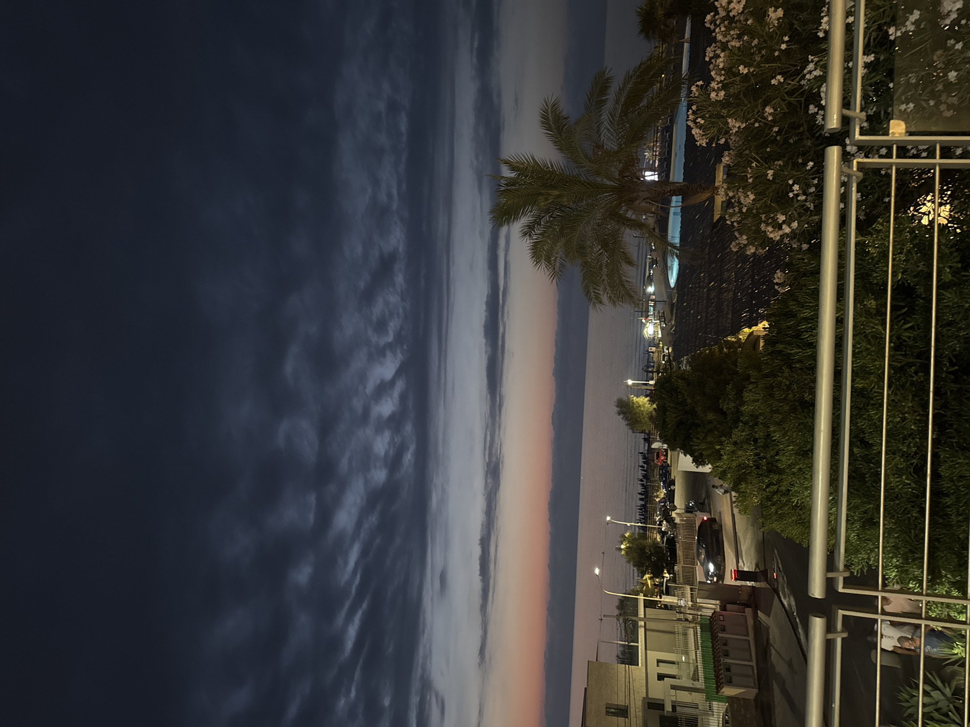 The image depicts a twilight scene in Capaci, Italy, overlooking the sea. Dark, striated clouds dominate the sky, with a band of pale orange and pink light visible on the horizon where the sky meets the water. The sea itself appears calm and dark. In the foreground, a metal railing runs across the bottom of the frame. Beyond the railing, lush green foliage is visible, interspersed with flowering bushes with small white blossoms. To the left, a building with white walls and dark window frames is partially visible. Further into the scene, a palm tree stands prominently, its fronds silhouetted against the darkening sky. Behind the palm tree, a swimming pool with illuminated blue water is visible. A beach with dark sand and rows of lounge chairs can be seen extending towards the sea. The scene is illuminated by artificial lights, including streetlights and lights from buildings, casting a warm glow on the surroundings. A few cars are parked along a road near the beach. In the lower left corner of the image, two individuals are partially visible behind the railing.