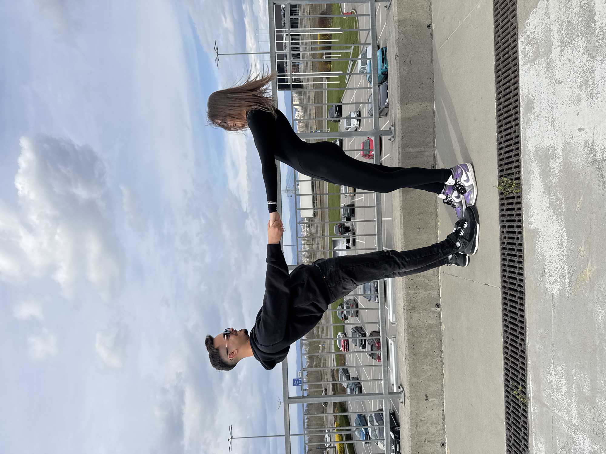 A man and a woman are holding hands, leaning over a railing on a high concrete structure. The woman is wearing a black jumpsuit and purple and white sneakers. Her hair is brown and blowing in the wind. The man is wearing a black hoodie, dark jeans, and black sneakers. He has sunglasses on. They are likely on a high point overlooking a parking lot and city area with multiple parked cars. In the background, there are buildings and overcast skies. The location is likely in the city of Sofia, Bulgaria.