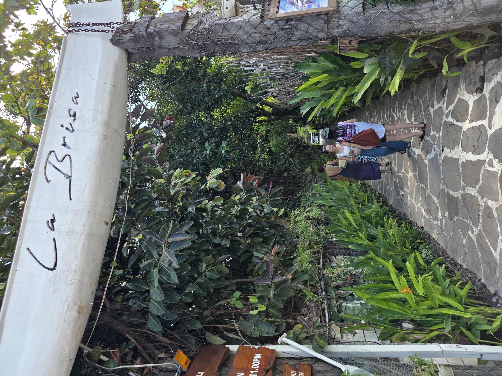 The image shows an entrance to a place called "La Brisa" in North Kuta, Indonesia. The entrance is an archway made of a white, weathered beam supported by thick wooden posts wrapped in netting. The words "La Brisa" are written in dark, stylized lettering on the beam. Lush green foliage and trees frame the entrance, with large, glossy leaves dominating the background.
A paved pathway, constructed from irregular, grey flagstones, leads through the archway and into the establishment. On the left side of the path, a white metal fence runs alongside a stone wall covered in ivy. Several wooden signs are attached to the fence, one with orange text stating "IN 11.00PM."
Three women are walking away from the camera on the pathway. The woman in the foreground on the right is wearing a white tank top, black shorts, and a baseball cap, carrying a red tote bag. To her left, a woman in a white tank top and blue pants smiles, also carrying a bag. Further ahead and to the left, another woman in a dark blue dress walks away.
The overall scene suggests a tropical, inviting atmosphere, possibly a restaurant or beach club, given the name and the abundant greenery. The lighting indicates it is daytime, with sunlight filtering through the trees.