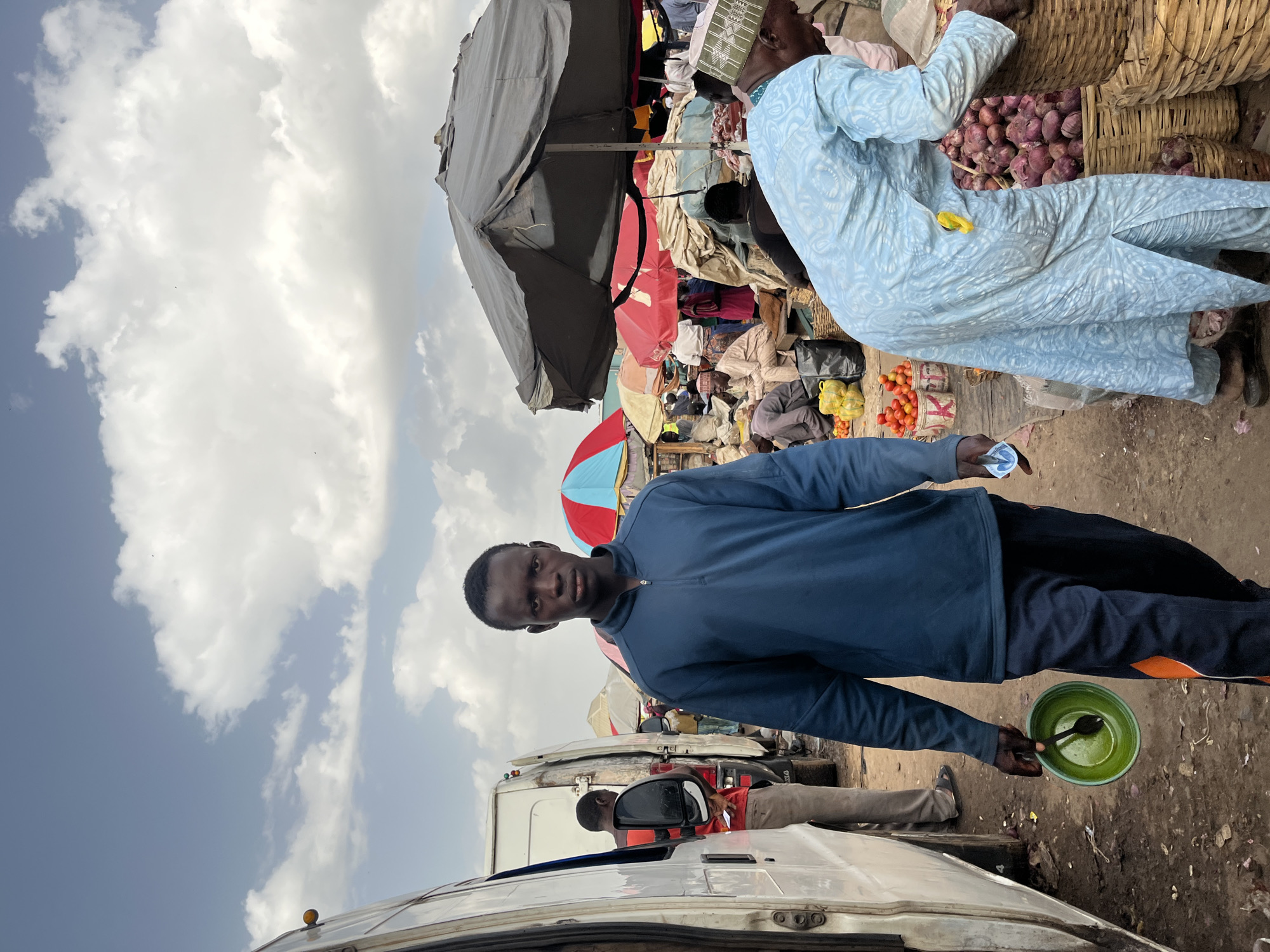 In Rafin Par, Nigeria, a young man stands in a bustling outdoor market, holding a green bowl with a spoon. He wears a dark blue quarter-zip sweater and dark pants with an orange stripe. Behind him, the market is alive with activity: several white vans are parked, and a man in a red and black striped shirt is visible near the side of one. Stalls display colorful umbrellas, with vendors and shoppers milling about amidst baskets of produce, including a large pile of onions in the foreground. The ground is dusty and uneven. The sky above is a mix of blue and scattered white clouds.