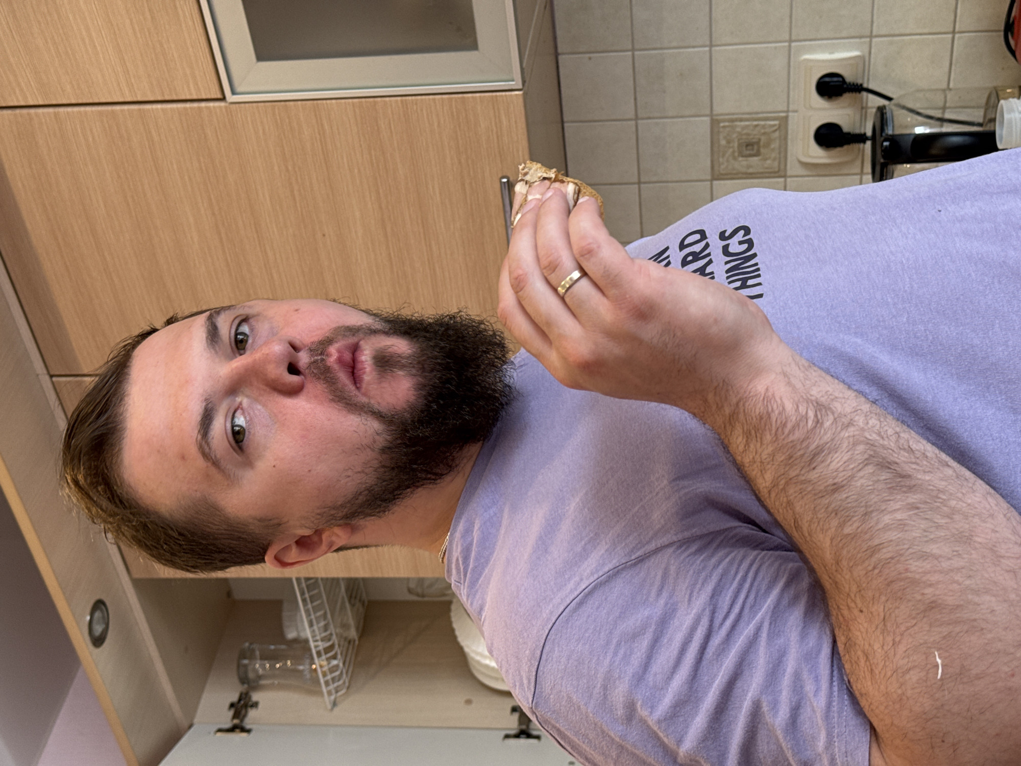 The image shows a close-up portrait of a man indoors, specifically in what appears to be a kitchen. The man, with short dark hair and a full beard, is looking directly at the camera with a slightly pursed-lip expression. He is wearing a light purple t-shirt, on which partial black text "ARD THINGS" is visible.
He holds a small, irregularly shaped piece of food, resembling a crusty piece of bread or pastry, in his right hand, which is raised near his mouth. A simple gold band ring is prominent on his right ring finger.
The background reveals a kitchen setting with modern, light-colored wooden cabinets. Above what appears to be a counter, there is a light-toned tiled backsplash with a subtle square pattern. Two white electrical outlets with black cords plugged into them are visible on the tiled wall. To the lower left, an open cabinet reveals a glass bottle and a white metal drying rack. The overall lighting suggests it is daytime. The man's posture and the food in his hand imply he is either about to eat or in the process of eating.