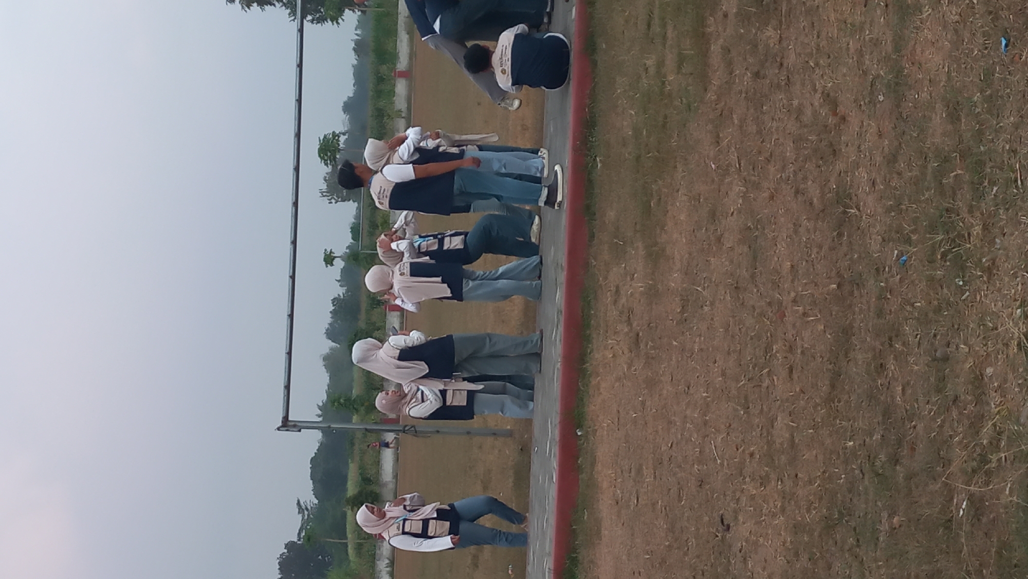 In Pare, Indonesia, a group of individuals are gathered on a grassy field, near a sports facility with a metal frame structure. The sky is overcast and the lighting is dim, suggesting it might be early morning or late afternoon. Most of the people are wearing matching vests over white shirts and jeans, with several of them wearing hijabs. They appear to be engaged in some kind of activity or event, possibly a team-building exercise or a class. One person is seated on the ground, while others are standing and interacting. The environment is mostly natural, with dry grass and a few trees visible in the background.