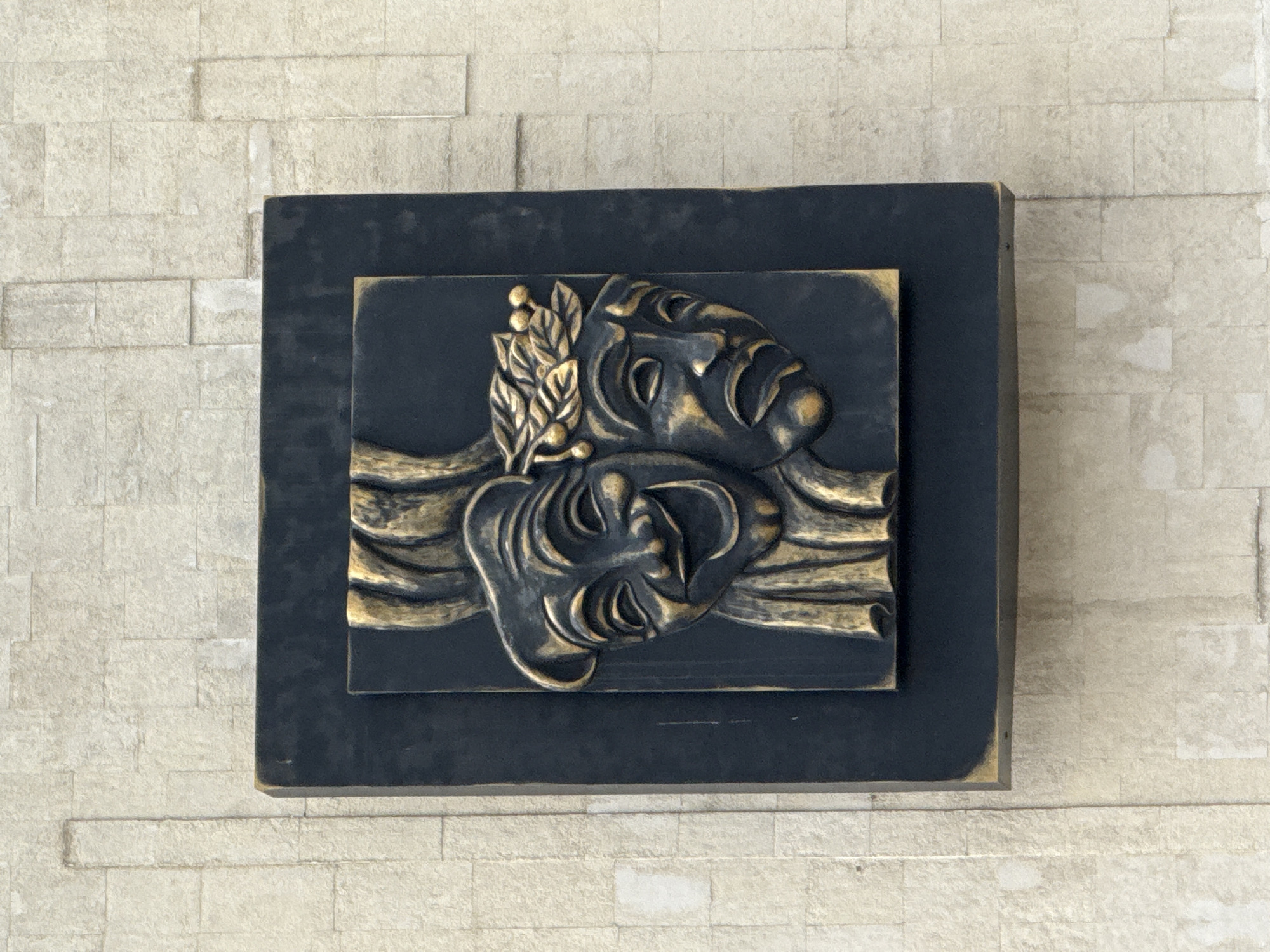 This image shows a bas-relief sculpture of two theater masks, one smiling and one frowning, mounted on a textured stone wall. The sculpture is dark with golden accents and is framed by a dark rectangular border. The wall is made of light-colored, rectangular stone blocks. This sculpture is located in Izhevsk, Russia, on the facade of the Udmurt State Academic Theater.