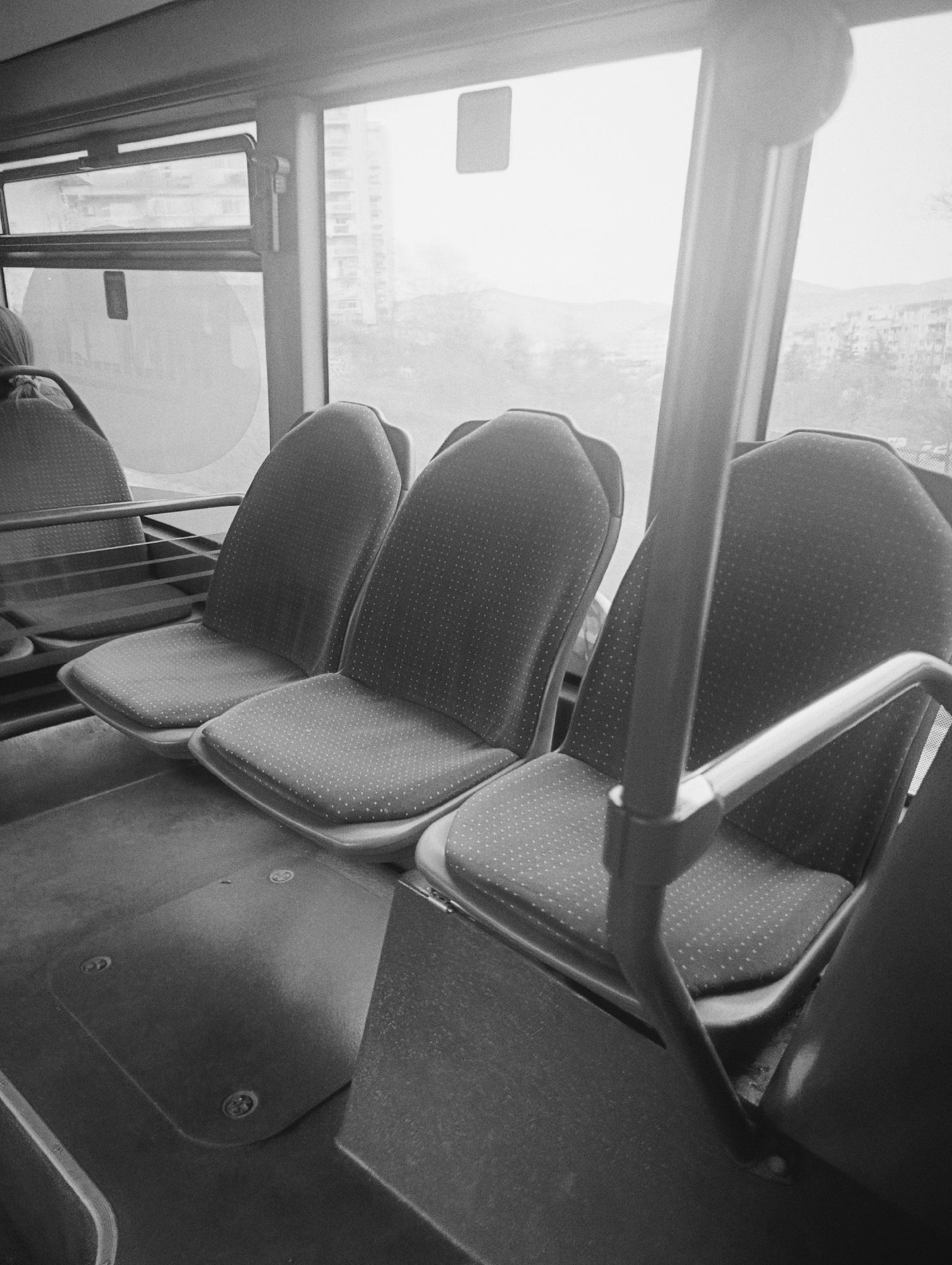 The image shows the interior of a public bus in Kardzhali, Bulgaria. Three unoccupied seats are visible in the foreground, upholstered in a fabric with a small, repeating dotted pattern. A vertical support pole and a horizontal handrail are prominent features near the front of the seats. Through the bus windows, buildings and a hillside are seen. The image is in black and white.