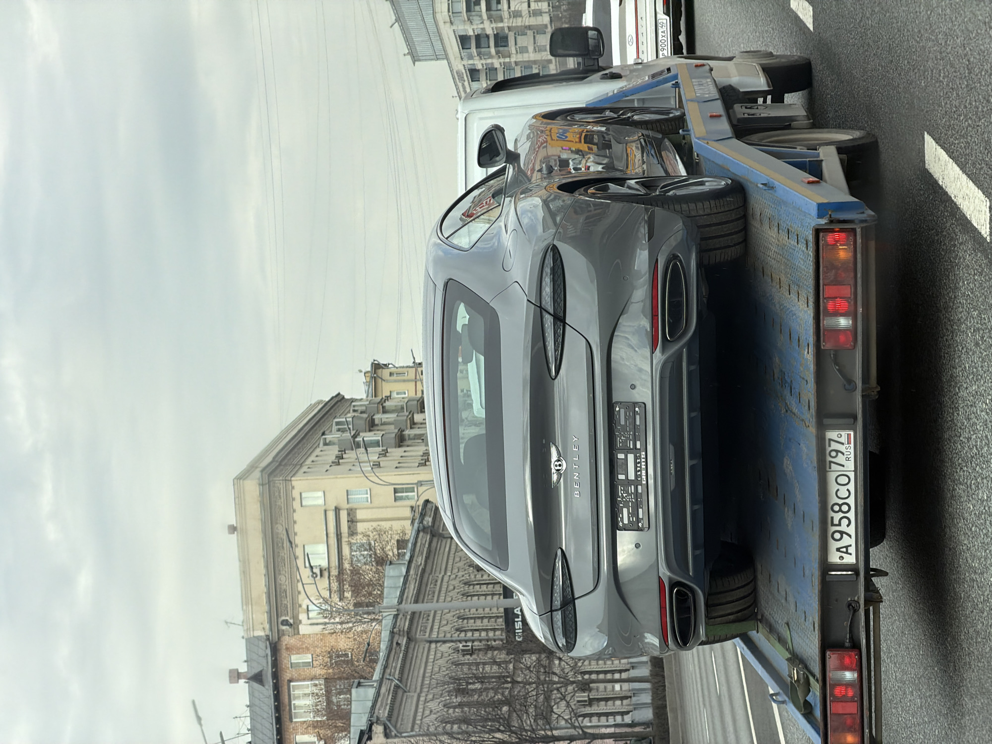 A gray Bentley Continental GT is being transported on a blue flatbed tow truck on a city street. The car's rear is facing the viewer, displaying the "Bentley" emblem and lettering, as well as the license plate. The tow truck's license plate reads "A958CO797 RUS". In the background, an older multi-story building with visible windows and architectural details lines the street. Overcast skies and a paved road surface are visible. The setting appears to be an urban area. The city is Moscow, Russia, as indicated by the license plate code "797 RUS".