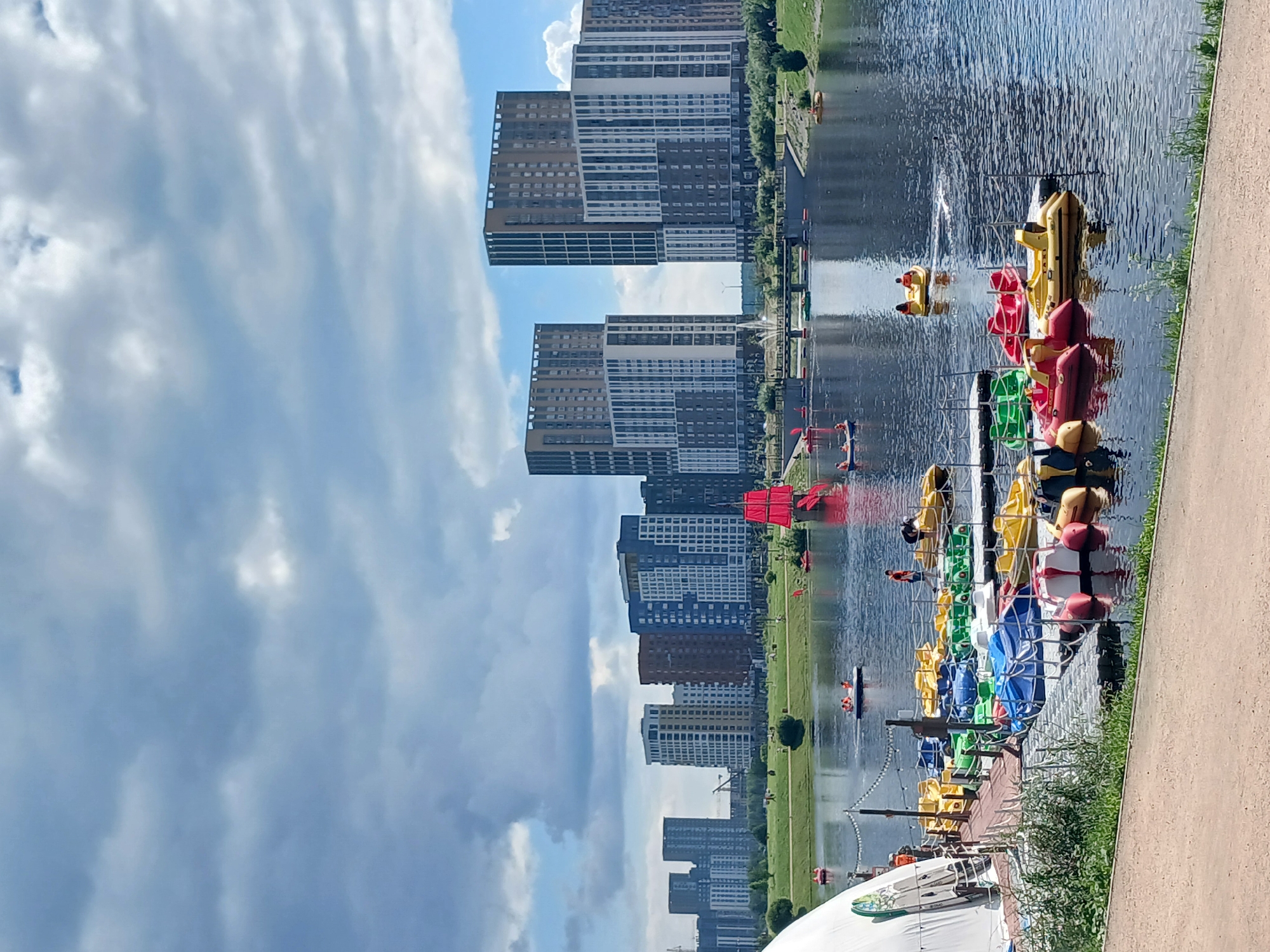 This is a photograph of a park with a lake in Yekaterinburg, Russia. The park is located in the area of Lake Shartash. In the foreground, a dock is filled with numerous colorful pedal boats, primarily yellow, green, and red. Several people are visible in and around the boats, some appearing to be operating them. The lake is calm and reflects the sky and the buildings. In the mid-ground, people are walking and relaxing on the grassy banks of the lake. A distinctive red-sailed replica ship is docked on the water. In the background, a modern city skyline is dominated by tall, multi-story residential buildings with contemporary architecture, some featuring balconies and large windows. The sky is partly cloudy with patches of blue visible. The overall scene depicts a recreational area within an urban environment.
