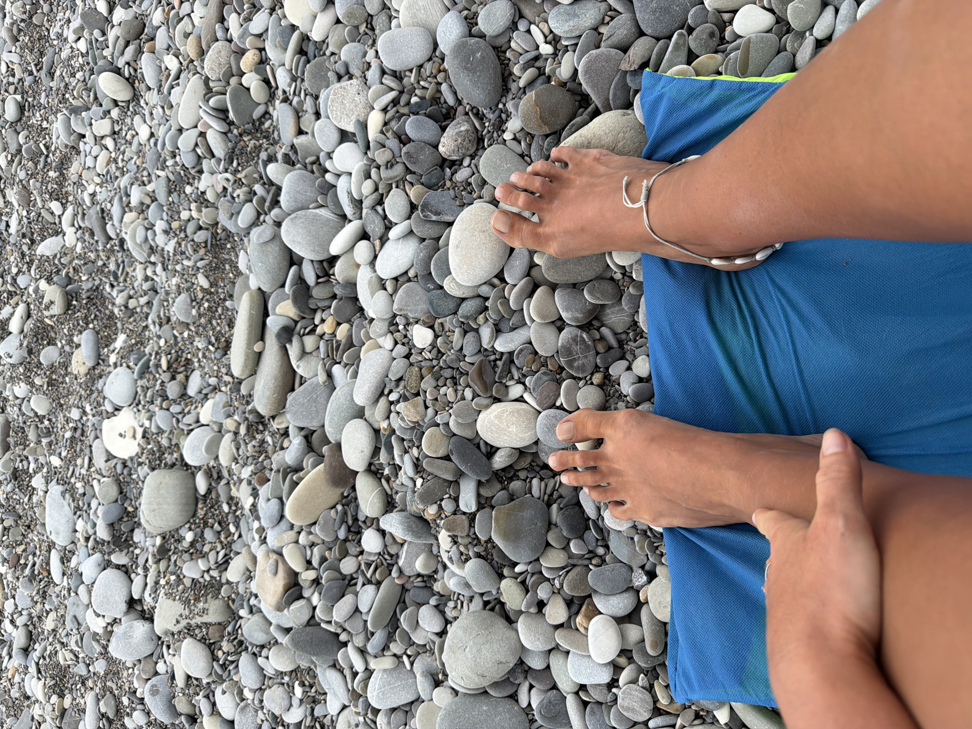 This image shows a close-up, overhead view of a person's feet resting on a blue towel on a pebble beach. The beach is covered in a variety of smooth, grey and white pebbles of different sizes, interspersed with darker sand. The person's feet are bare, and one foot is adorned with a delicate, silver-colored anklet featuring small white shells. The skin tone suggests a sunny environment. The towel is a vibrant blue and appears slightly damp in places, hinting at recent contact with water or a humid atmosphere.
The scene is likely set during daylight hours, given the clear visibility and absence of shadows that would indicate twilight or night. The focus on the feet and the beach suggests a leisurely activity, possibly relaxation or a break from swimming, common in a coastal resort town like Sochi, Russia. The overall impression is one of calm and simple enjoyment of a natural, seaside setting. There is no visible text in the image.