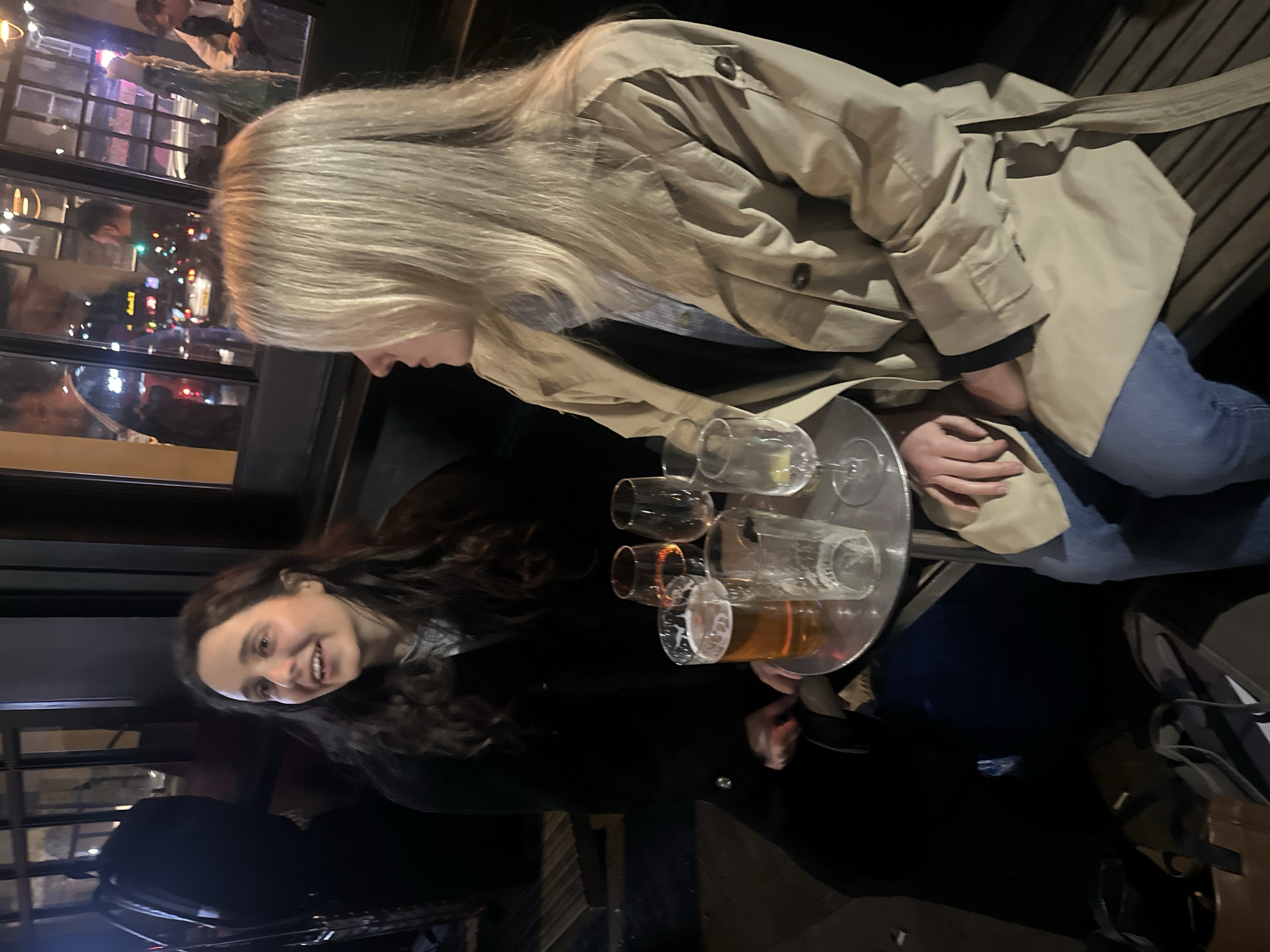 Two women are sitting outside a restaurant or bar in London, United Kingdom. The woman to the left has long, wavy brown hair and is smiling. The woman to the right has long blonde hair and is wearing a trench coat and jeans. They are sitting at a table with a metal tray holding several glasses, one of which contains beer. The background includes reflections of city lights and other people in the restaurant's windows.