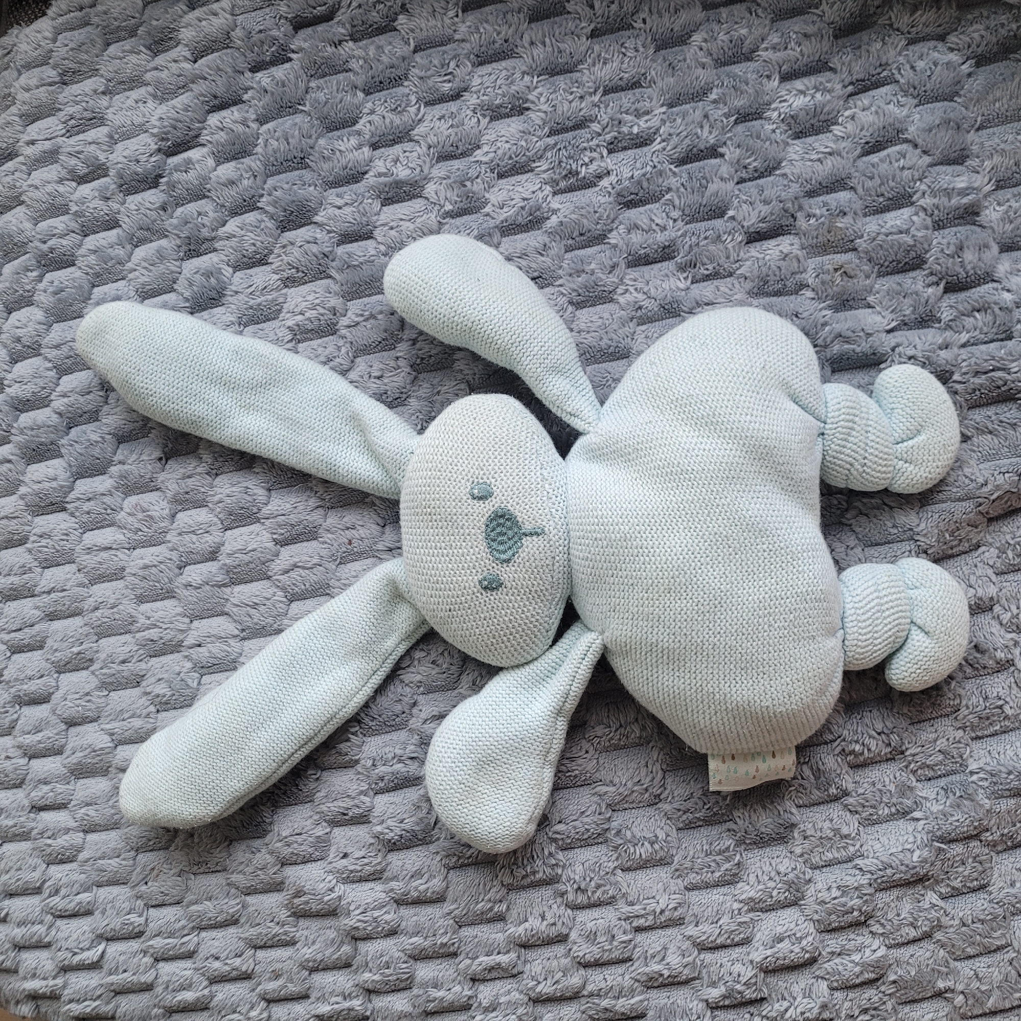 A light blue knitted bunny stuffed animal is lying on its back on a textured gray blanket. The bunny has long ears, a round head with embroidered eyes and nose, and short arms and legs. The blanket has a raised pattern of interlocking hexagonal shapes. There is a small tag with a pattern of colorful dots and leaves on the left side of the bunny's body. The image is taken from a top-down perspective. The location in Jūrmala, Latvia is not discernible from the image.