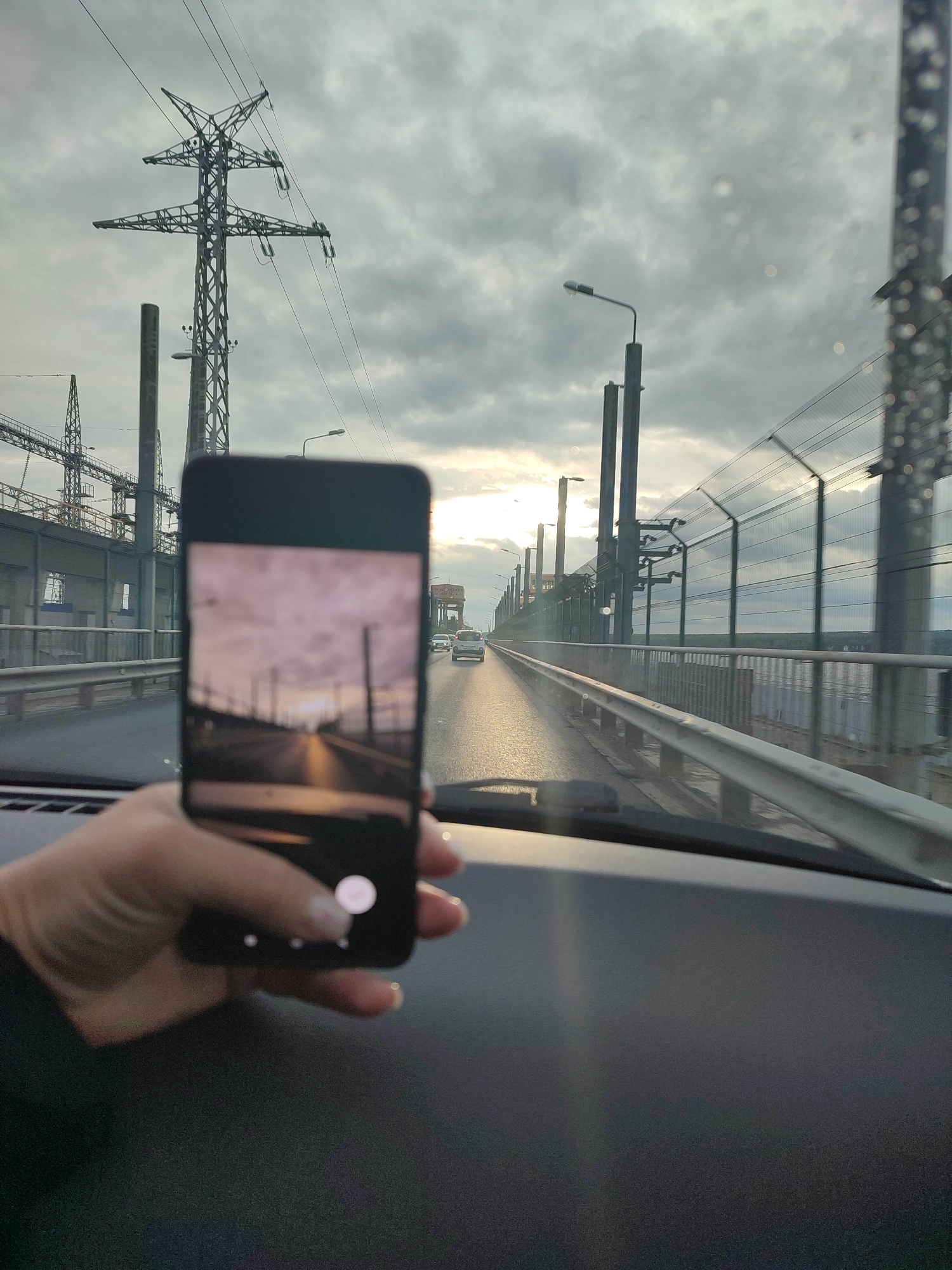 The image captures a first-person perspective from inside a vehicle, looking through a windshield that shows visible water droplets. A person's left hand, with manicured nails, holds a black smartphone vertically in the foreground, positioned over the vehicle's dark dashboard. The phone screen displays a photograph of a road scene, illuminated by a low sun creating a bright reflection on the asphalt and casting a warm glow on the clouds. The phone's interface shows a white circular button and three smaller dots below the image.
The environment outside the vehicle depicts an elevated road or bridge, likely over water, with a multi-lane highway extending into the distance where several cars are visible. To the right of the road, a metal guardrail runs parallel, followed by a tall, transparent security fence with a diamond mesh pattern. Beyond the fence, a body of water is visible, with distant land or trees on the horizon. Along the road, numerous streetlights and tall, slender utility poles are present. To the left of the road, a large lattice-work power pylon with star-shaped cross arms supports multiple electrical lines, adjacent to an industrial complex featuring various metal structures and additional power lines. The sky is overcast with grey clouds, but the sun is low on the horizon, breaking through the clouds on the right side of the image, creating a bright glare and lens flare effects. The overall scene suggests either dawn or dusk. The location is in the city of Unknown, Unknown.