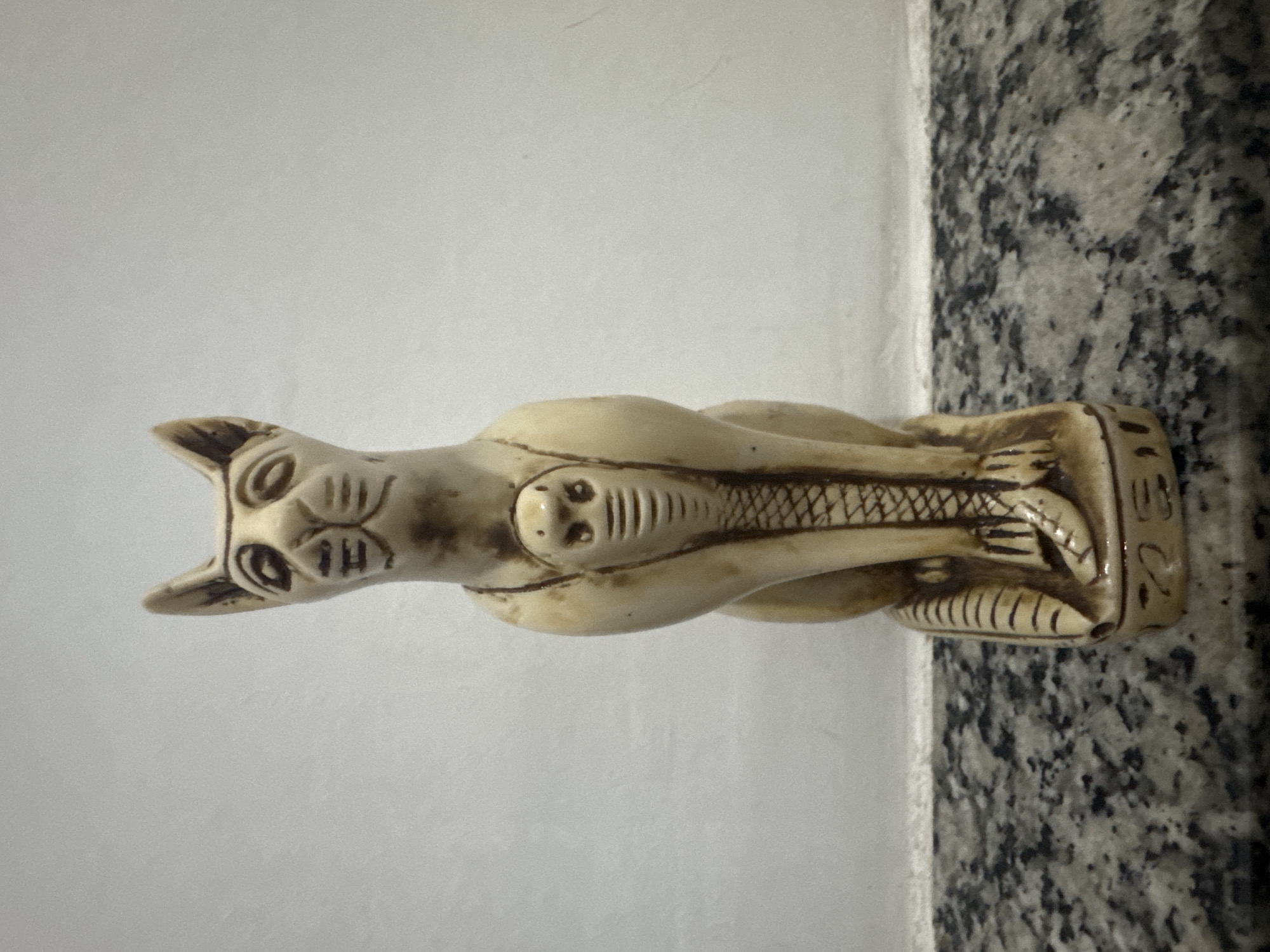 A light-colored, Egyptian-style statuette of a cat with a cobra coiled around its chest is centered against a white wall and positioned on a speckled grey countertop. The cat statuette is upright, with its head facing forward and its ears pointed upwards. Dark lines are etched onto the face to depict eyes, nose, and whiskers. The cobra statuette has its hood flared, and it is wrapped around the chest of the cat. The base of the cat statuette has some faded hieroglyphic markings. The countertop exhibits a mottled pattern of grey and black specks, with some lighter grey areas. The wall behind the statuette is plain and white, with some faint textural variations. The image was taken in Niterói, Brazil.