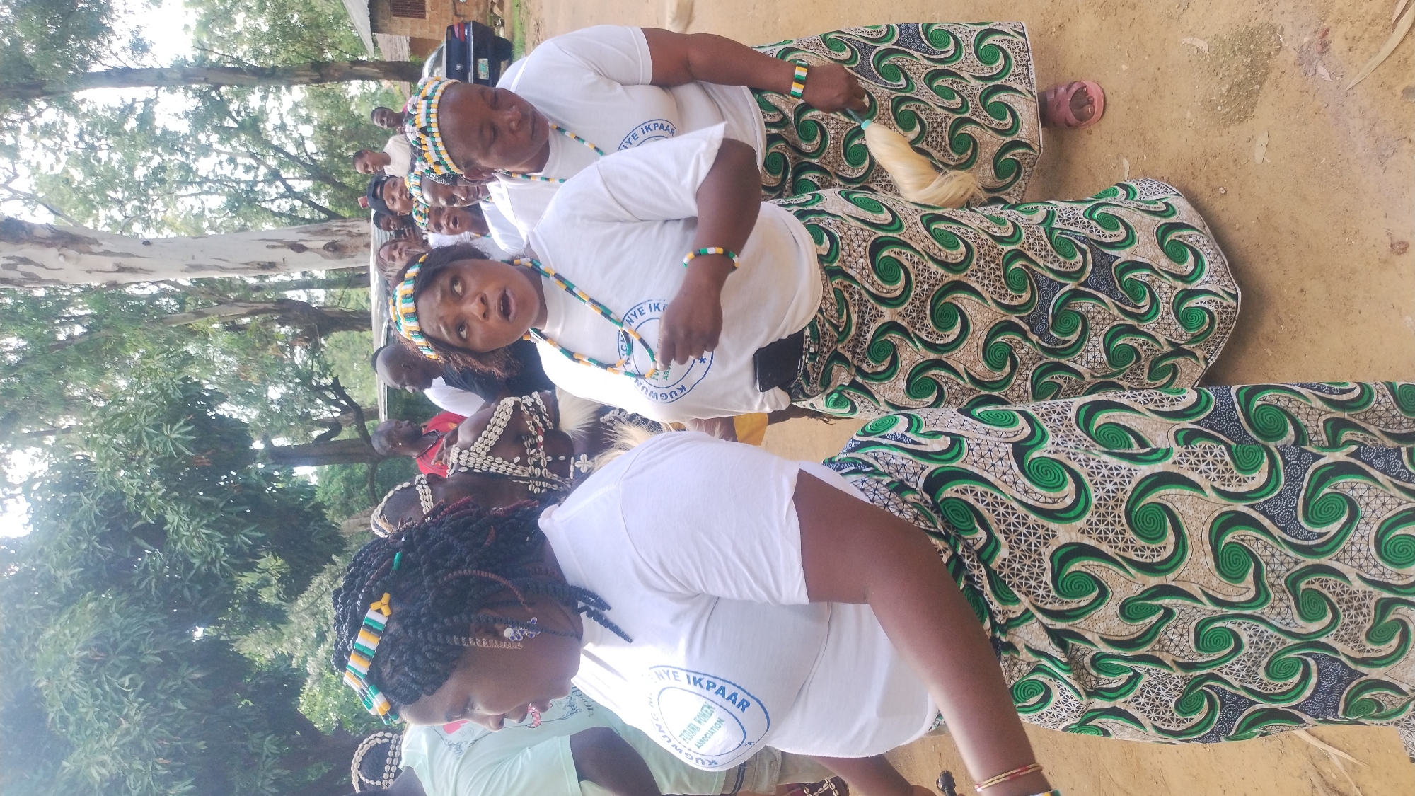 In a gathering in Jos, Nigeria, a group of women, adorned in traditional attire, are seen in motion. The women are wearing white t-shirts with blue circular logos and colorful headbands featuring yellow, green, and white geometric patterns. Their skirts are made of fabric with a distinctive pattern of swirling green shapes on a tan background. Some women are wearing beaded necklaces and bracelets in similar colors to their headbands. In the background, trees provide a natural backdrop, and a building and a car are partially visible, suggesting an outdoor setting within a semi-urban or rural area. The overall impression is one of a cultural event or celebration.