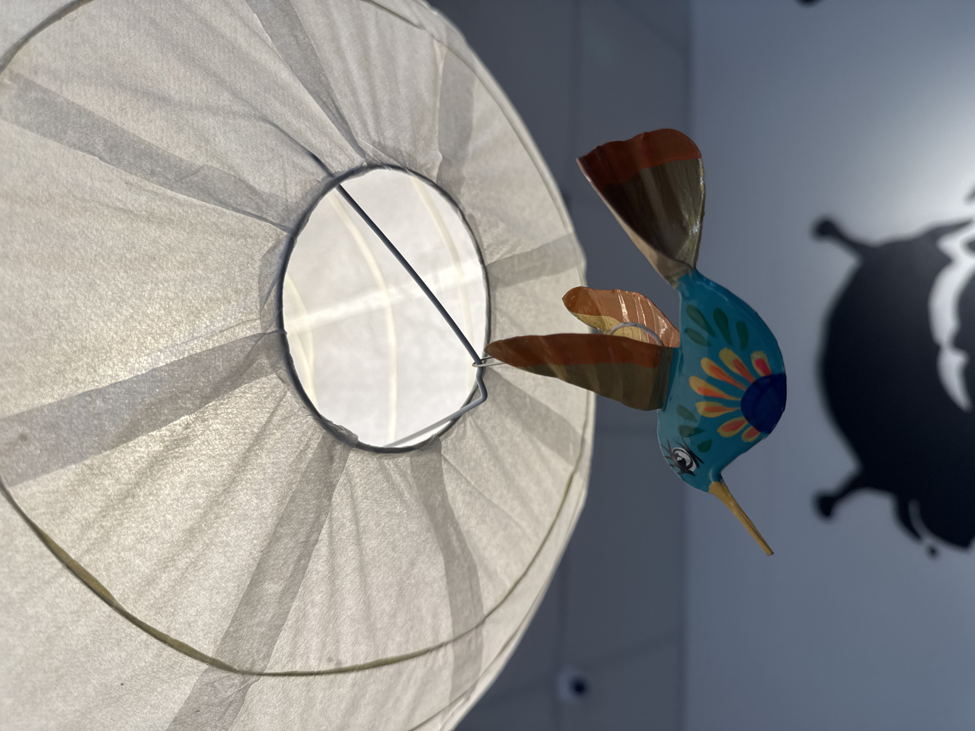 A colorful, stylized hummingbird decoration hangs from a wire beneath a large, white, translucent paper lantern. The hummingbird is predominantly teal with orange and yellow accents on its wings and tail. It has a long yellow beak and detailed painted eyes. The lantern is lit from within, casting a soft glow and creating shadows on the wall behind it. The background is a neutral, light-colored wall, and a faint shadow of what appears to be a decorative element or object is visible on the lower portion of the wall. The scene appears to be indoors, possibly in a shop or home. The city of Kremenchuk, Ukraine is not identifiable from the visual information provided.