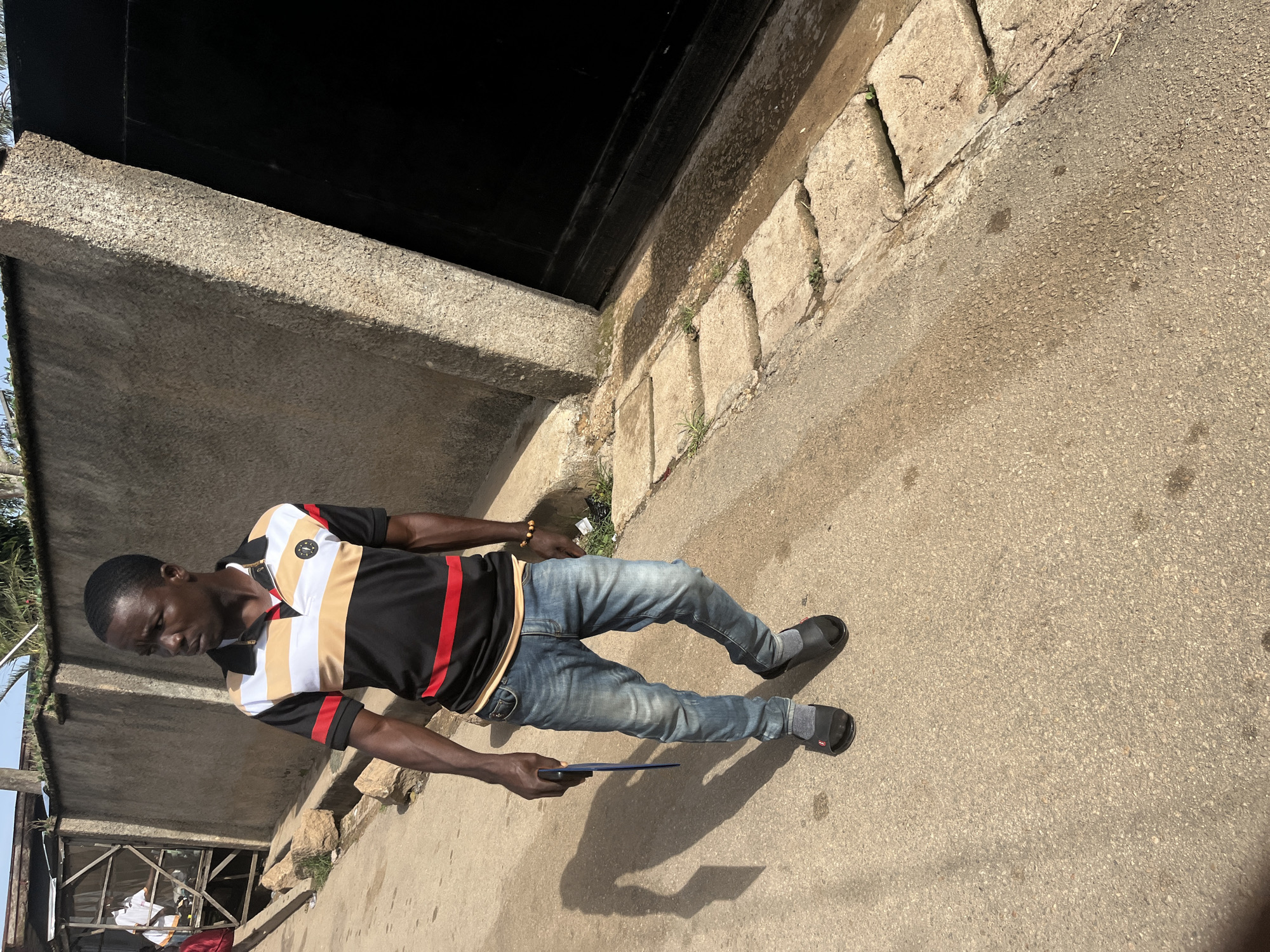 In Jos, Nigeria, a man walks on a paved road adjacent to a rough concrete wall and a large black metal gate. He is wearing a black polo shirt with white and gold stripes, blue jeans, and black sandals with grey socks. He holds a blue object in his right hand. The man is looking to his left, and his shadow is cast on the road behind him. In the background, to the left, a wooden structure with people is visible. The environment appears to be outdoors, likely in a residential or commercial area, with hints of vegetation growing in cracks in the pavement.