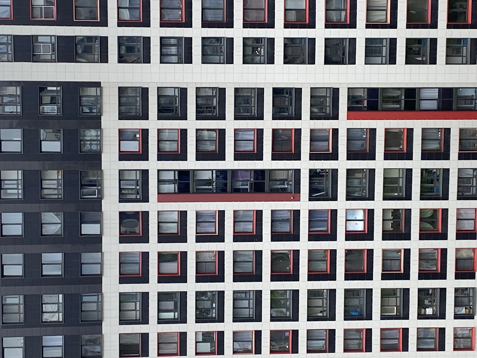 The image shows an exterior view of a modern building with numerous square windows. The facade is divided into two distinct sections: a section on the left with black window frames and a section on the right with white window frames. Some windows have red accents around the frames, especially in the right section. The building appears to be located in an urban area, but the exact location (city) is not identifiable. The windows reflect varying levels of light and possible interior contents, indicating they are not all vacant. There are no visible people, but the condition of the windows and the presence of possible items inside suggest the building is occupied.